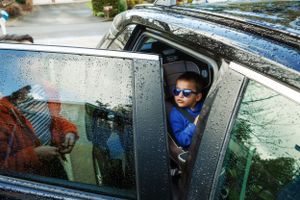 Someone wearing sunglasses peers out from a car window on a rainy day with water droplets visible on the glass.