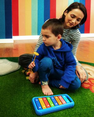 Two people sit on green carpeting with a colorful xylophone toy while surrounded by rainbow striped walls.