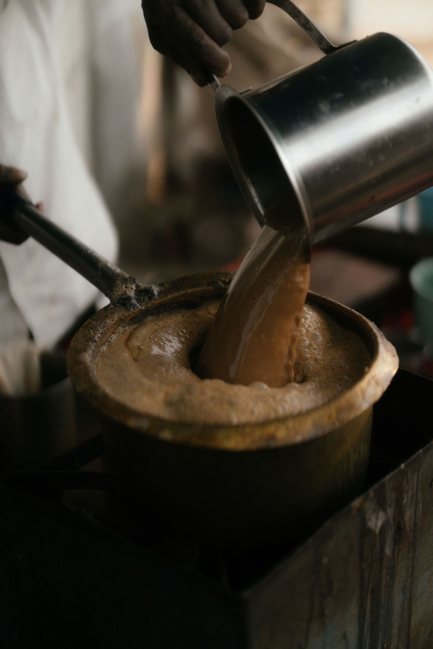 Dark liquid being poured from a metal pitcher into a rustic wooden vessel in moody lighting.