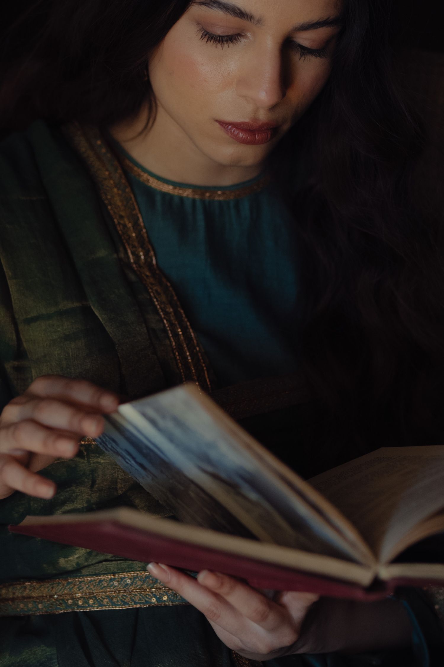 Figure in teal and green traditional clothing reads book in dramatic lighting.