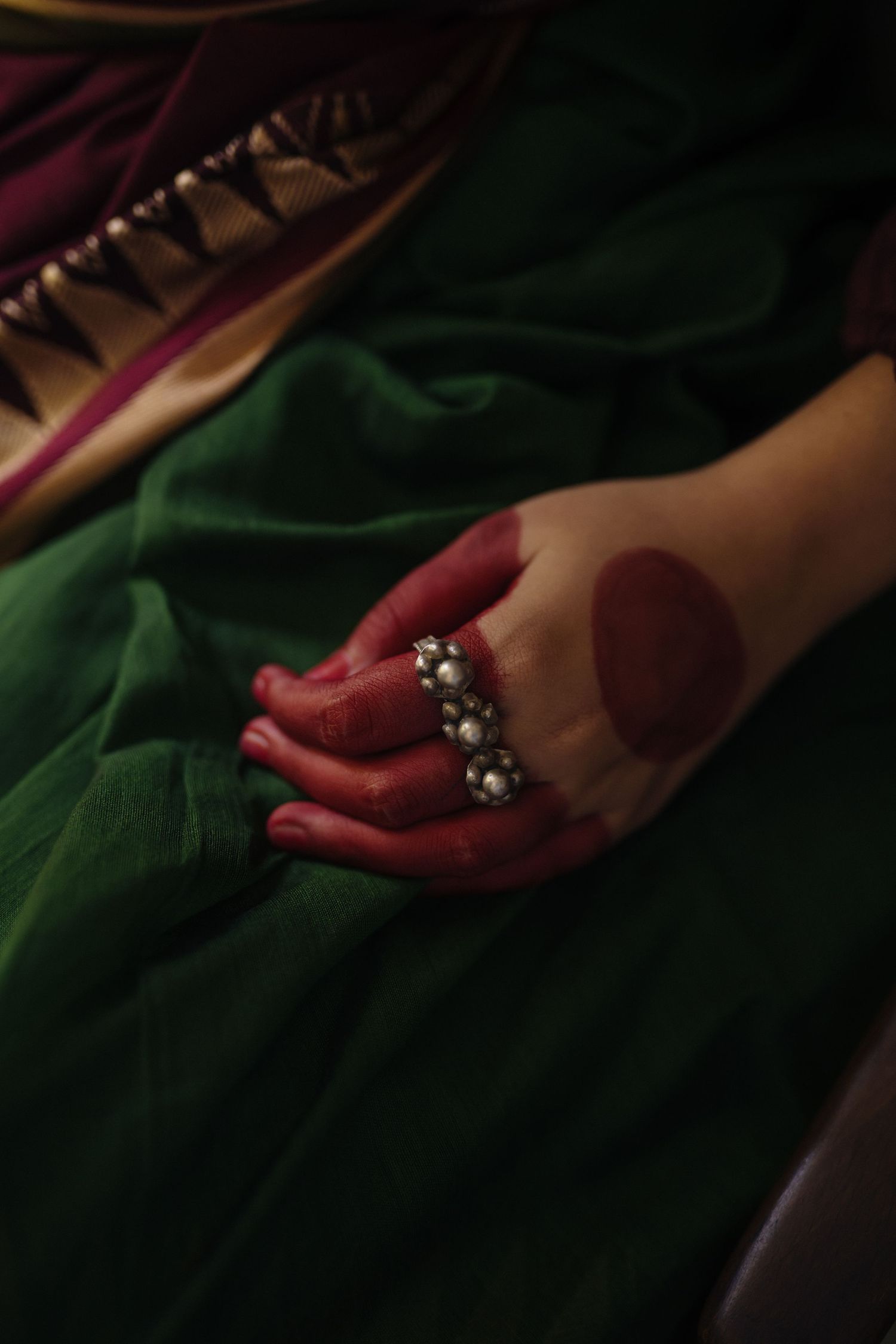 Red-stained hand wearing an ornate ring against green fabric in dramatic lighting.