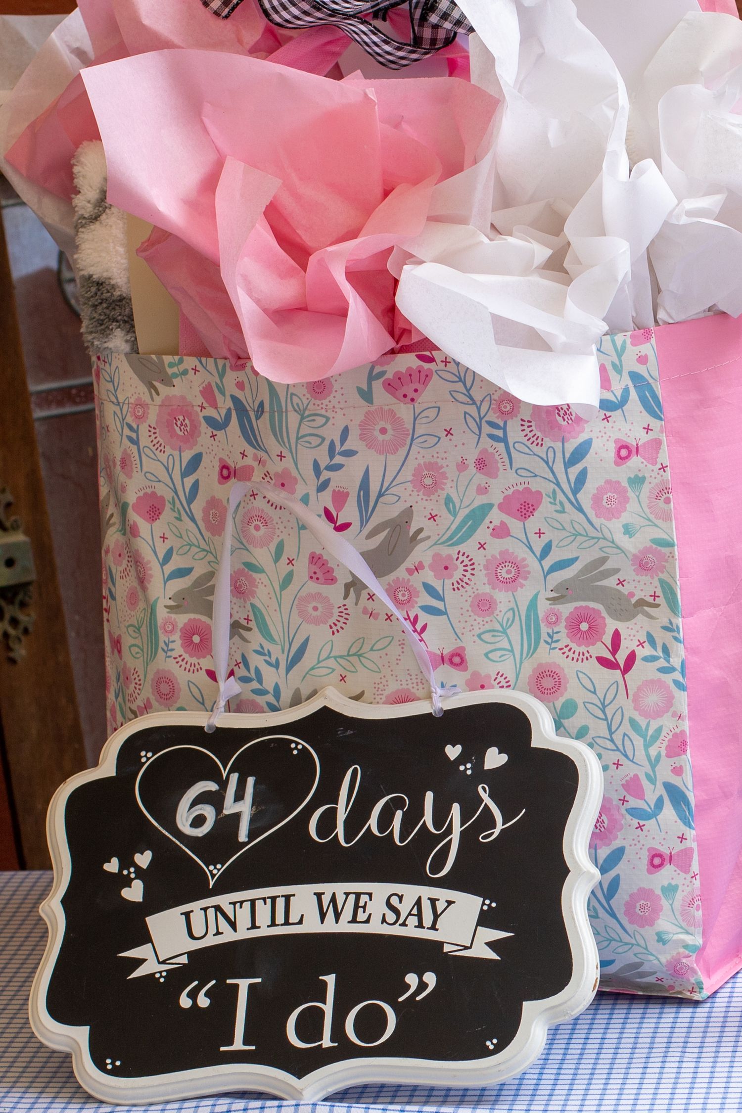 A chalkboard wedding countdown sign showing '64 days until I do' next to gift bags with pink tissue paper and floral print.