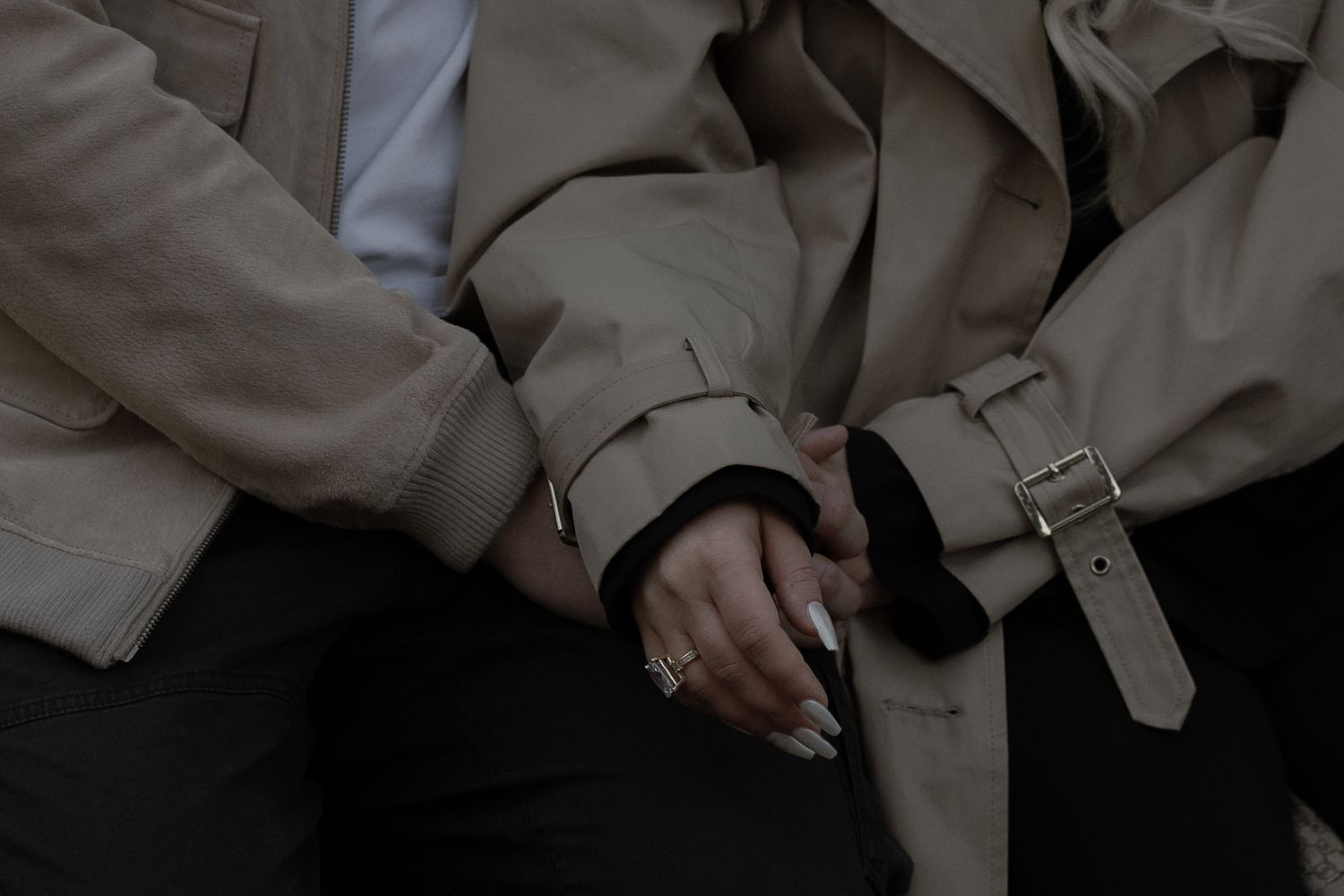 Close-up view of hands wearing a beige trench coat with buckled sleeves against dark clothing.