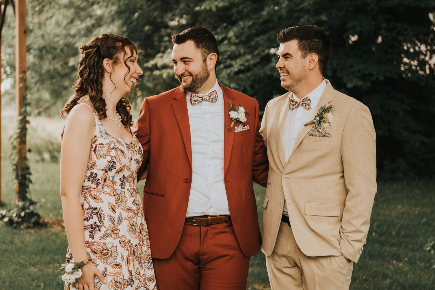 Three people sharing a joyful moment outdoors, wearing formal attire in rust red, tan, and floral print.