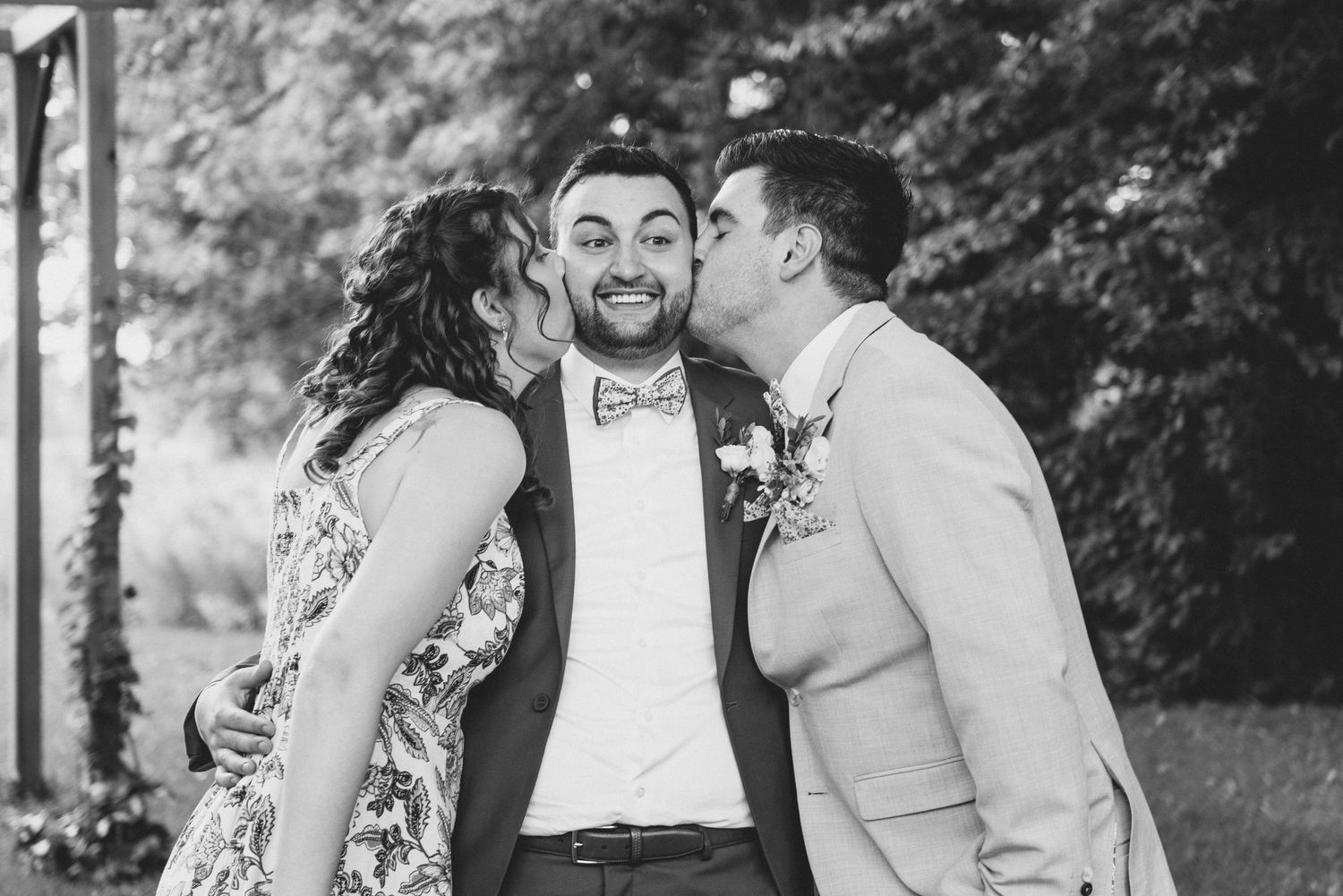 Black and white photo showing three stylishly dressed people sharing a joyful moment outdoors with kisses on cheeks.