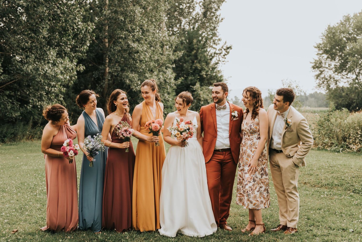 Wedding party stands outdoors in colorful earth-toned attire featuring rust and mustard bridesmaid dresses and beige suits.