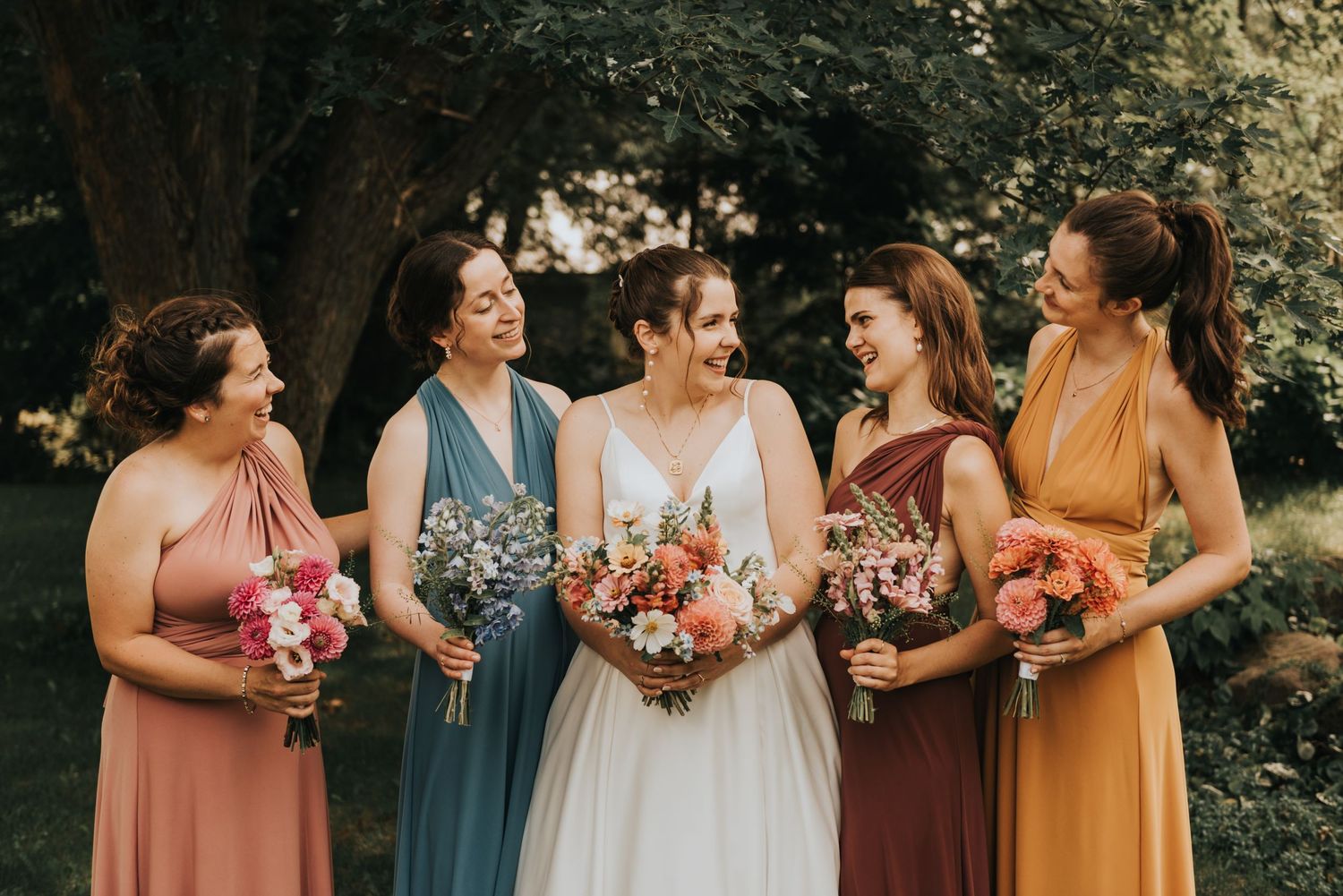 Wedding party of bridesmaids in coral, blue and rust colored dresses share candid moment outdoors.