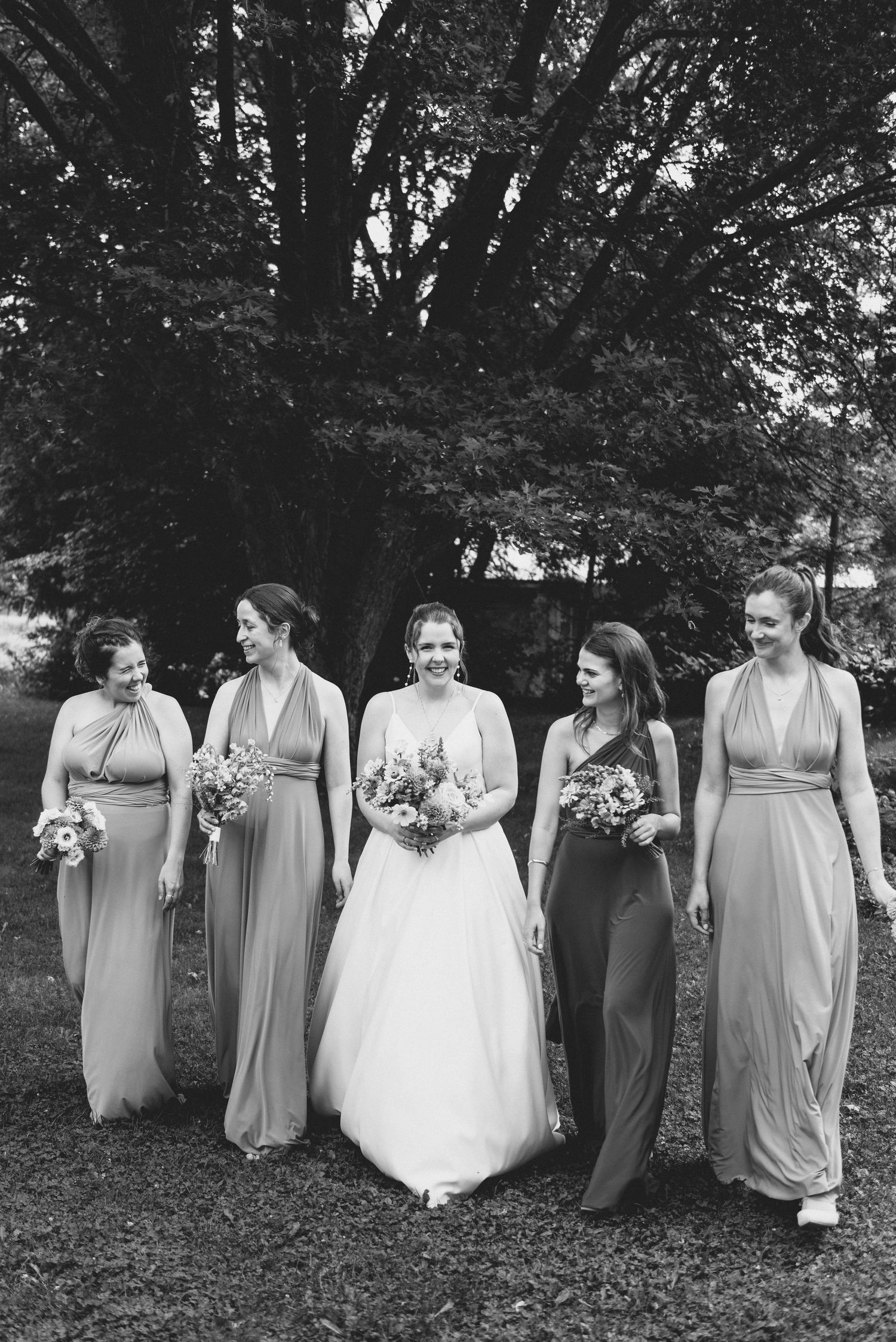 A wedding party in long dresses poses together outdoors under a large tree in a black and white photograph.