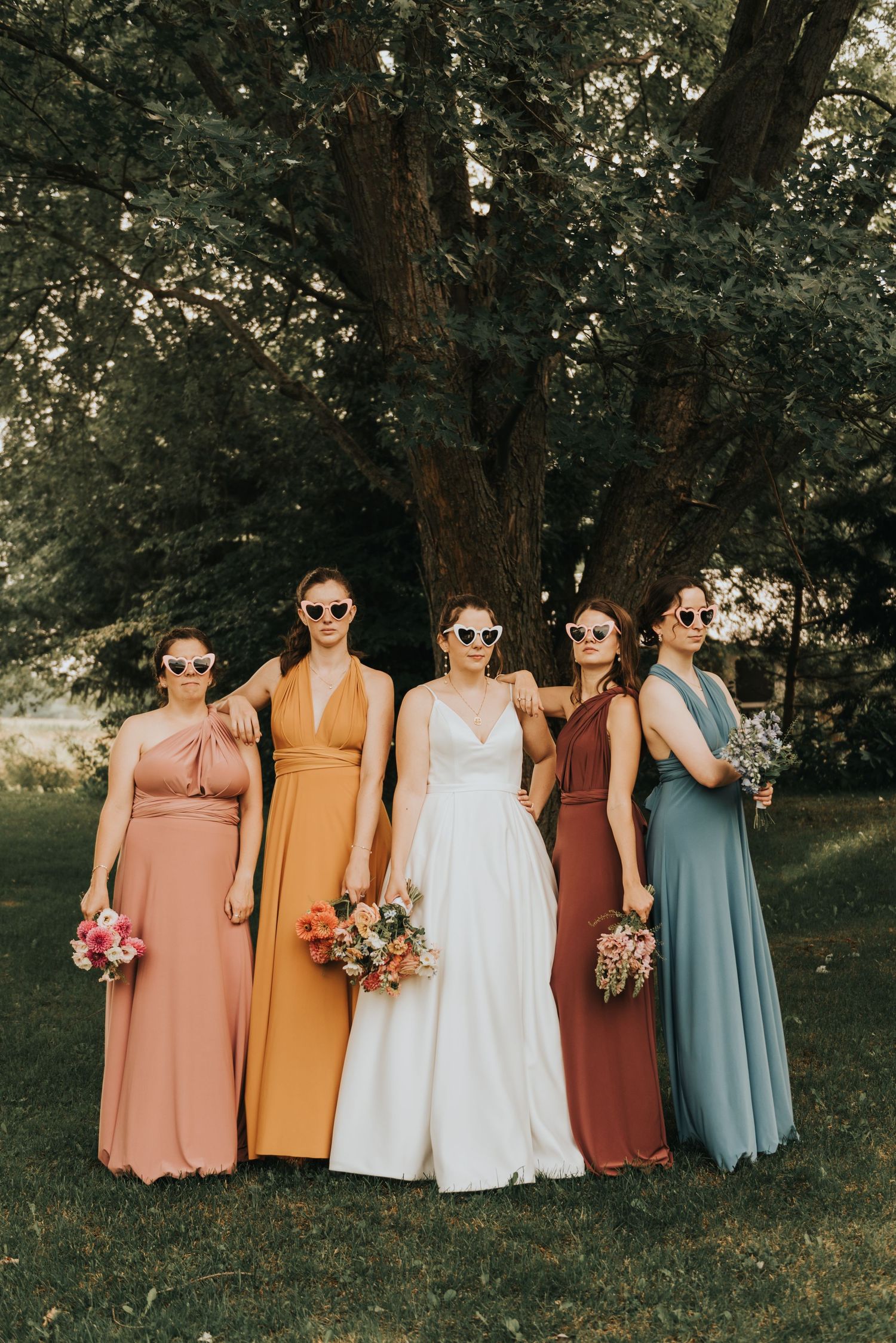 Bridal party poses outdoors in coordinating dresses of peach, yellow, white, rust and sage green colors.