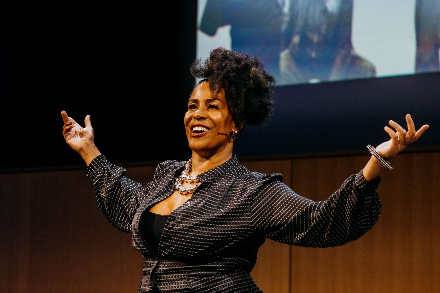 Animated speaker in polka dot dress gestures enthusiastically with outstretched arms on stage during presentation.