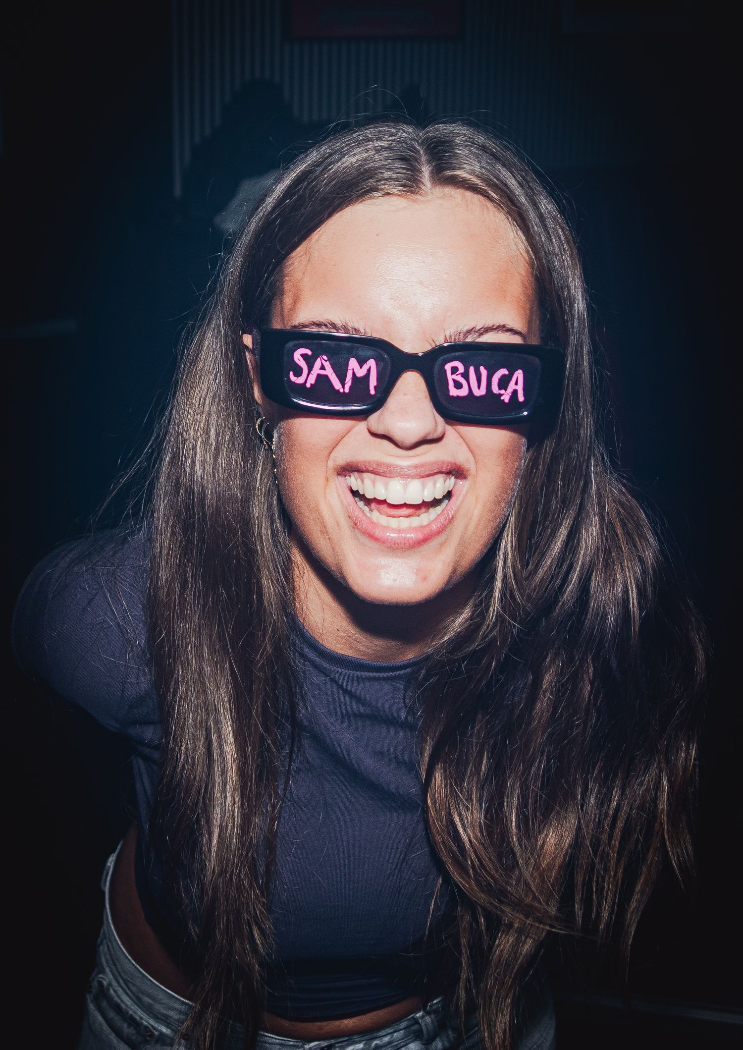Person wearing sunglasses with text "SAM BUCA" written on them smiles broadly against dark background.