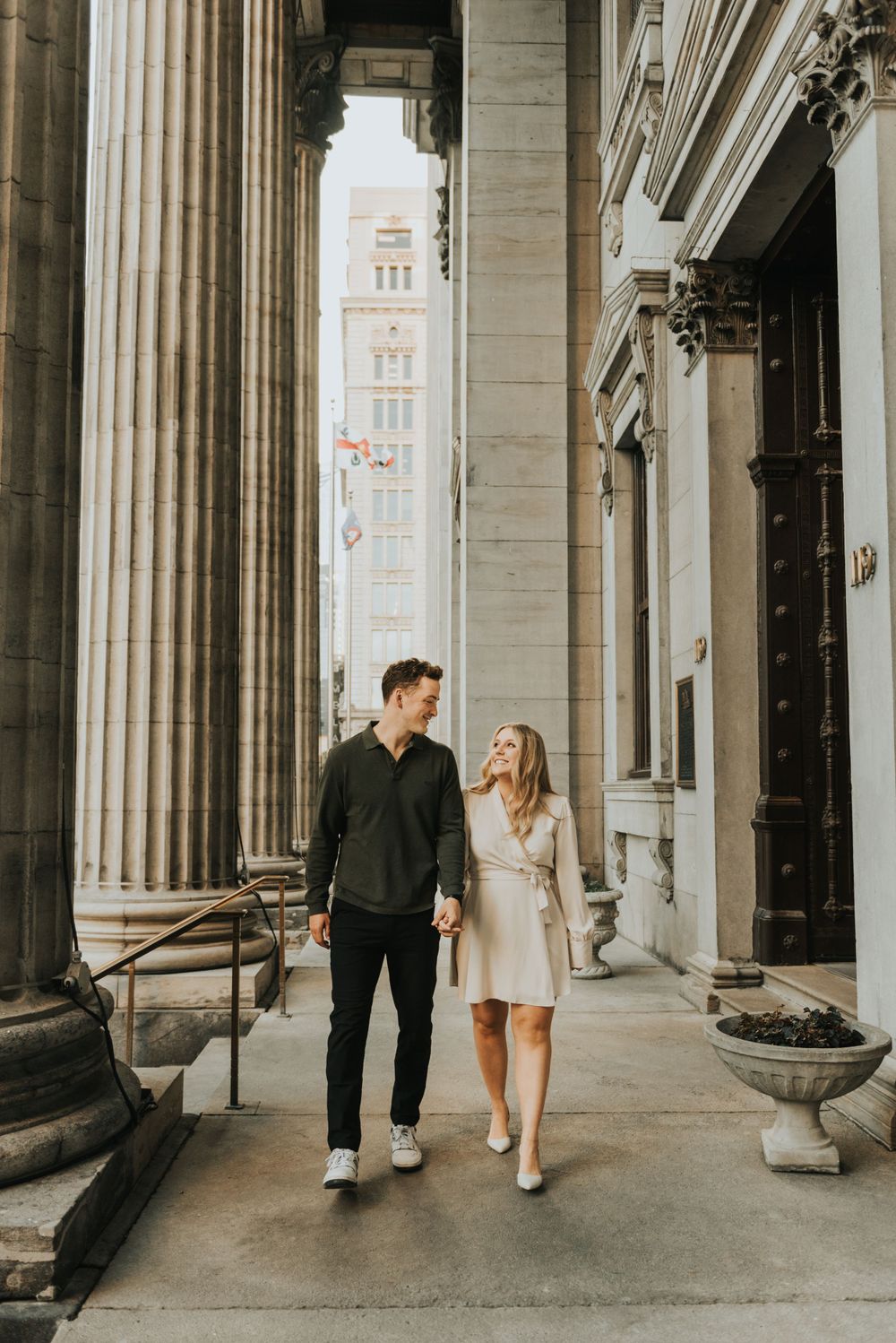 A couple strolls together between classic stone columns of a grand historic building with an ornate tower visible in background.