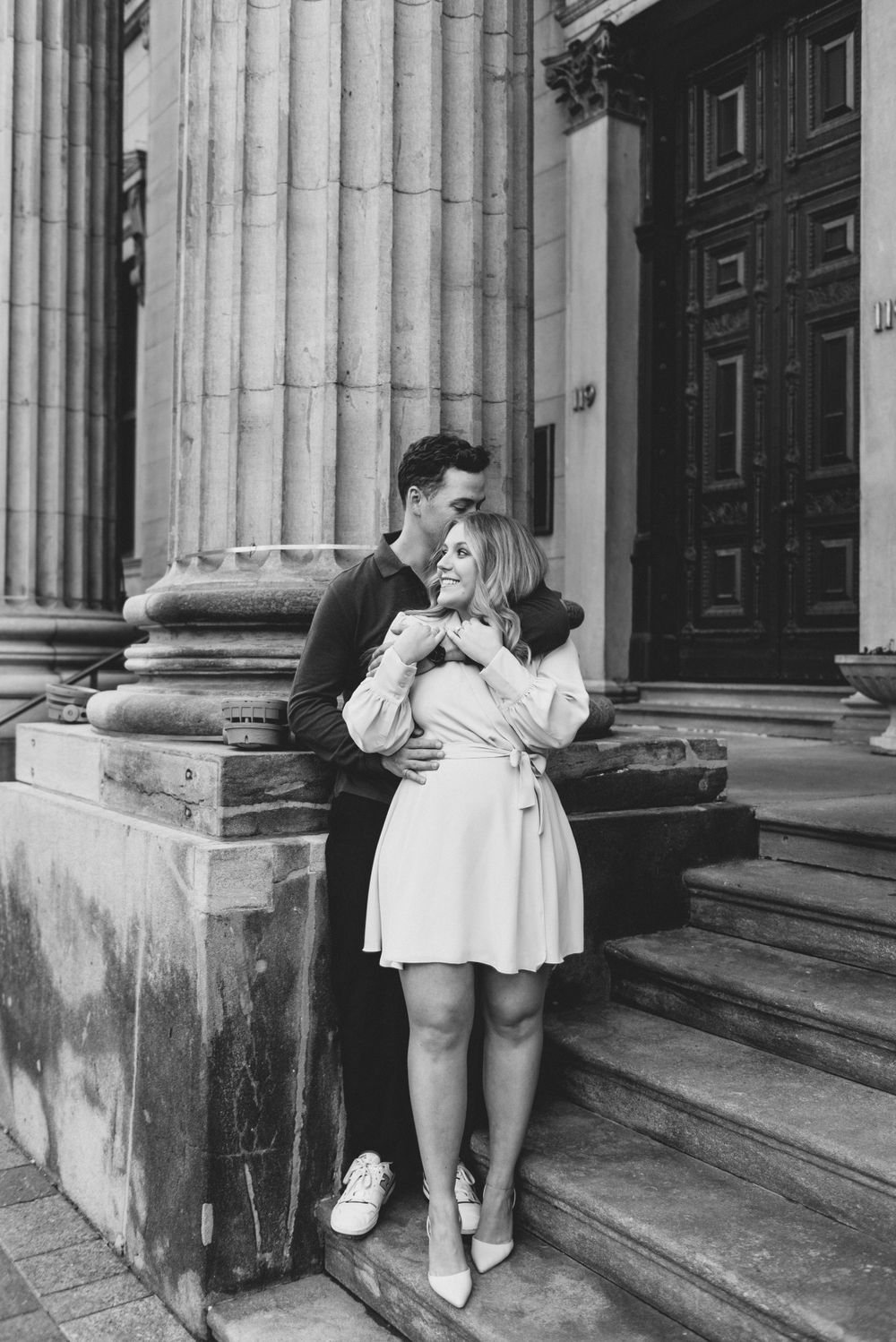A romantic couple in black and white embraces on stone steps near classical columns outside a historic building.