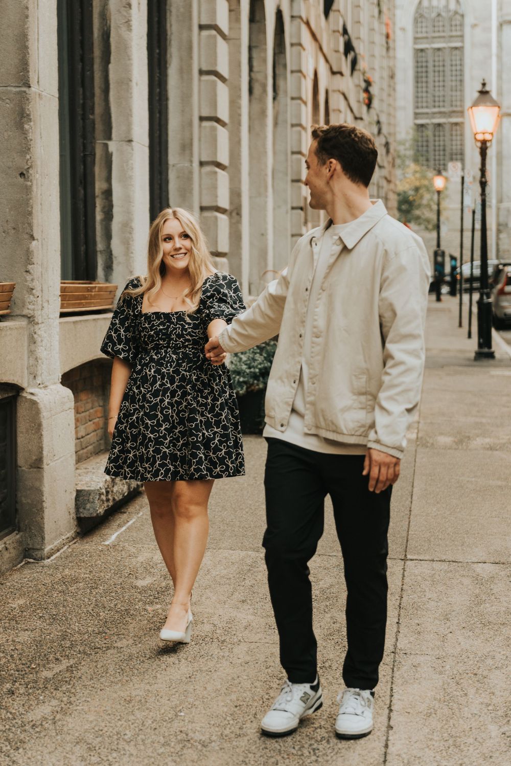 A couple walking together on a city sidewalk with vintage architecture and street lamps in the background.