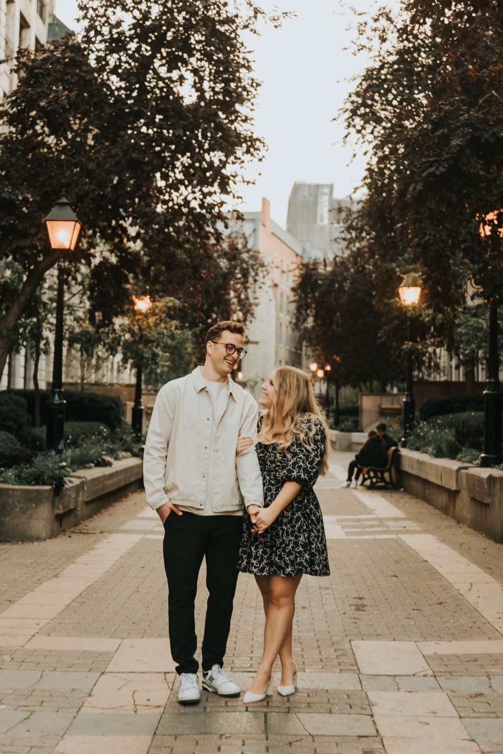 A romantic couple strolls down a lamp-lit urban pathway at dusk with buildings and trees in the background.