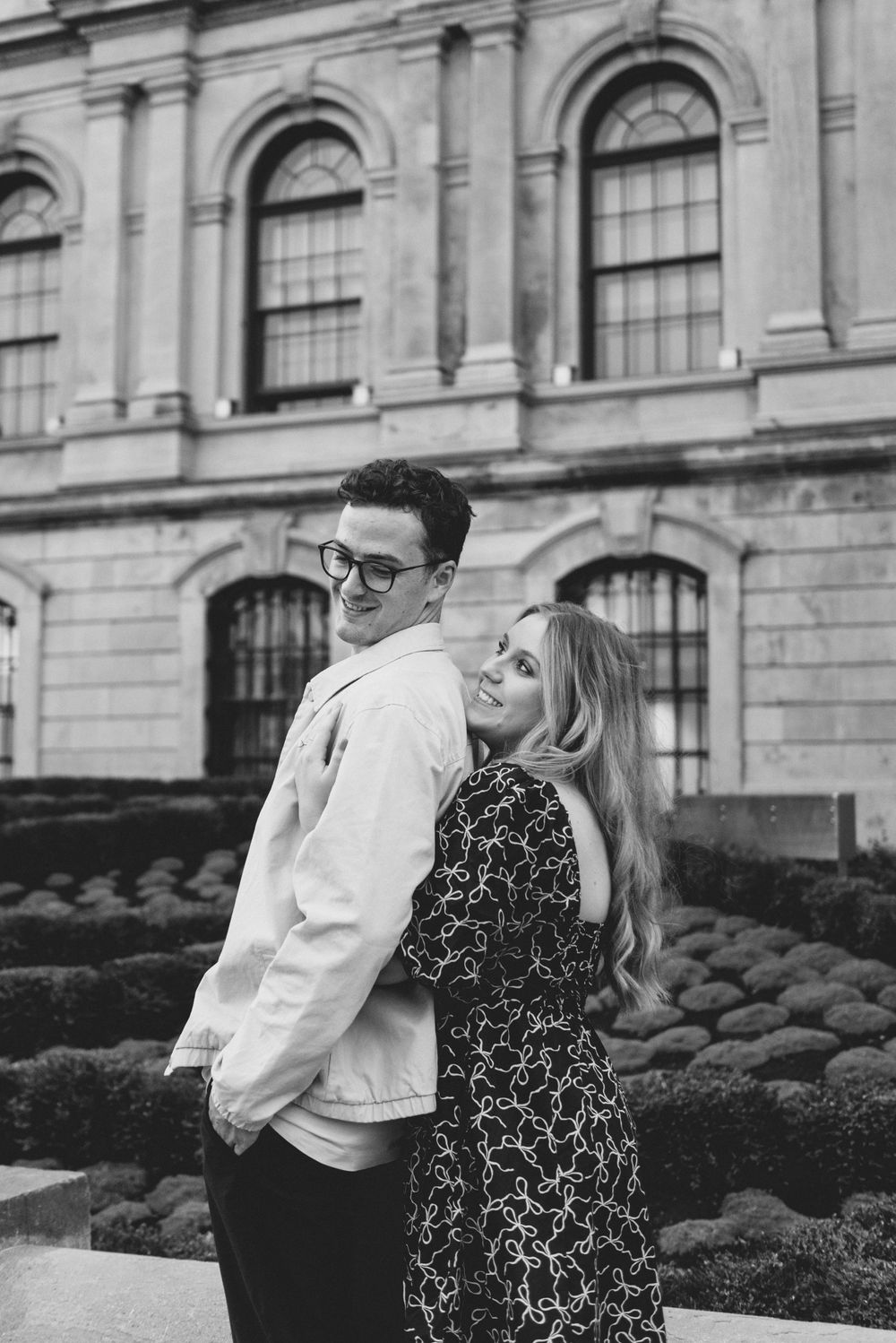 Black and white romantic portrait of a couple posing together in front of a historic stone building with arched windows.