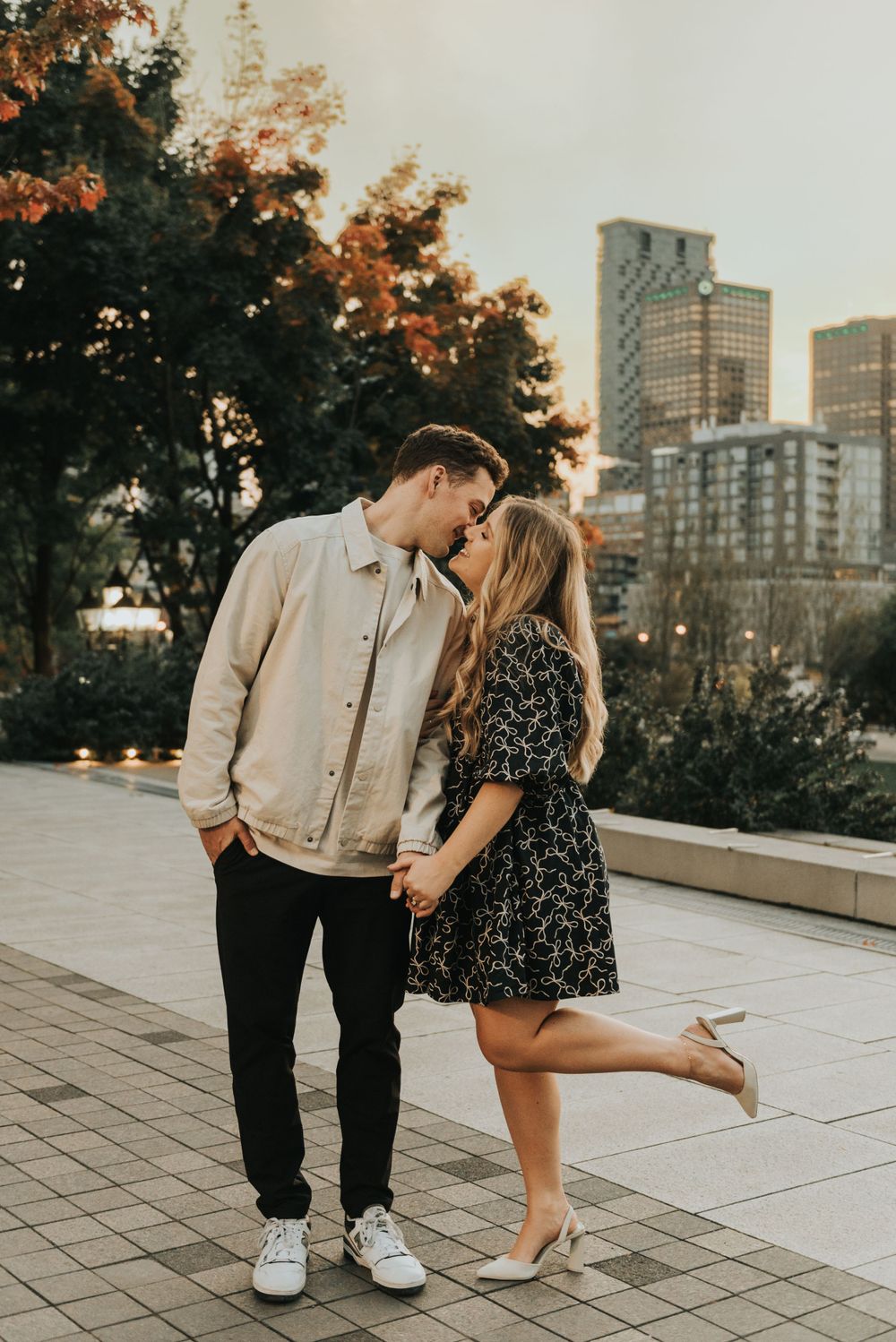 Romantic couple sharing a tender moment at sunset in front of modern city skyline with autumn trees in background.