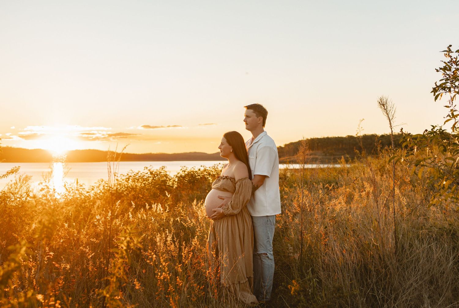 Couple embraces tenderly in golden sunlight among wild grasses and brush during outdoor photo session.