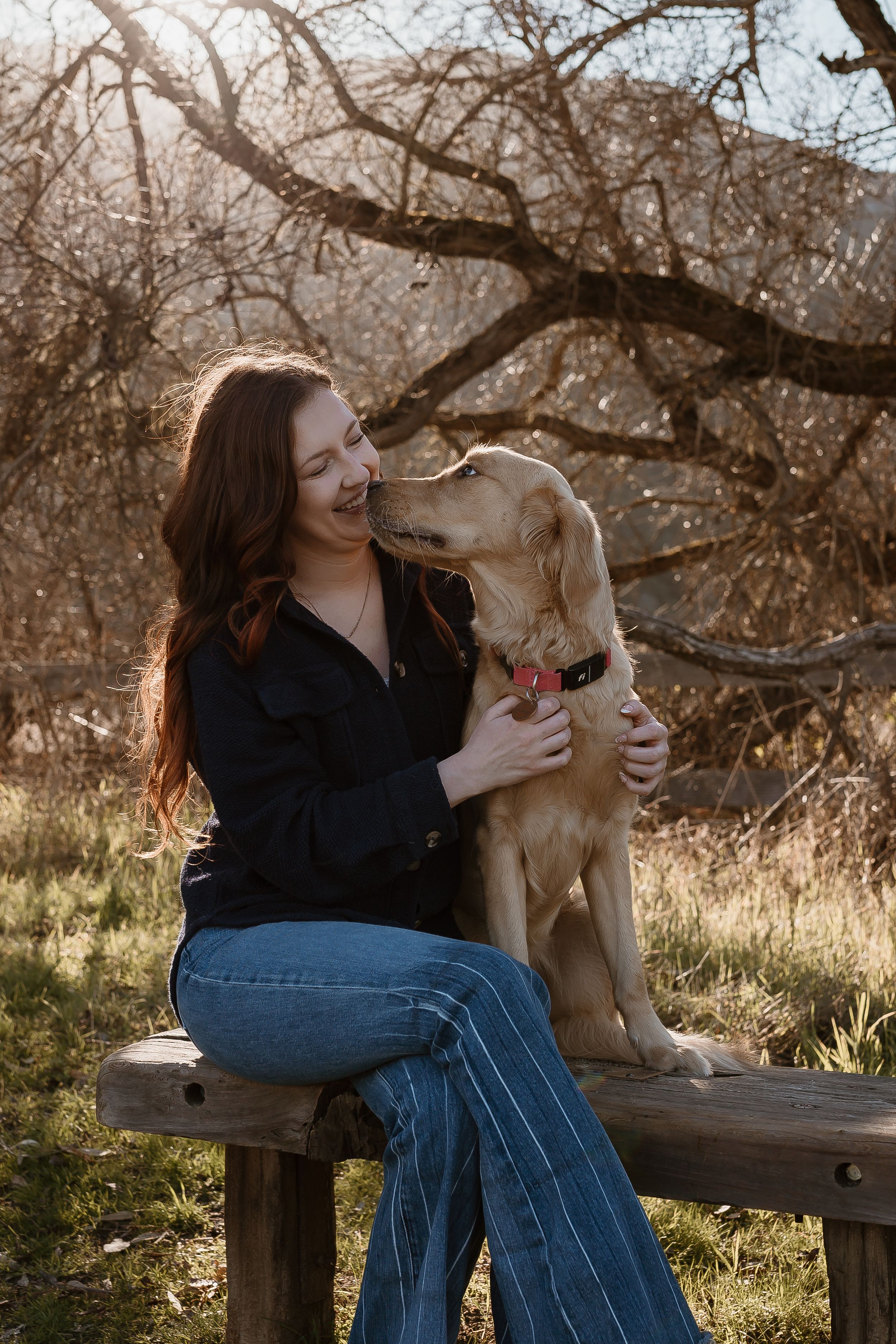 Woman in dark sweater and jeans sits on wooden bench sharing tender moment with golden retriever dog in autumn setting.