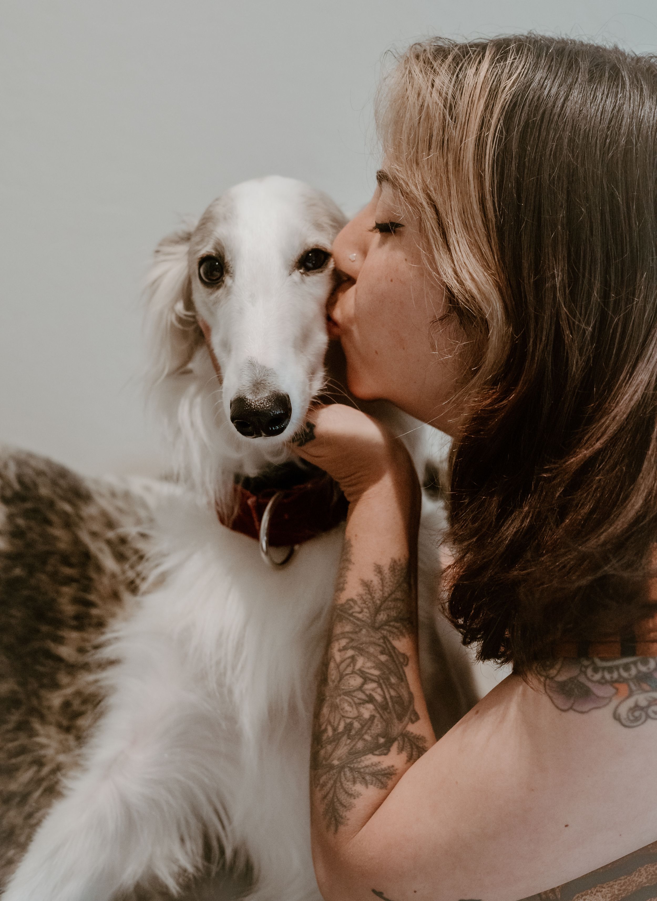 A person with visible tattoos gives a sweet kiss to their white borzoi dog in an intimate portrait.