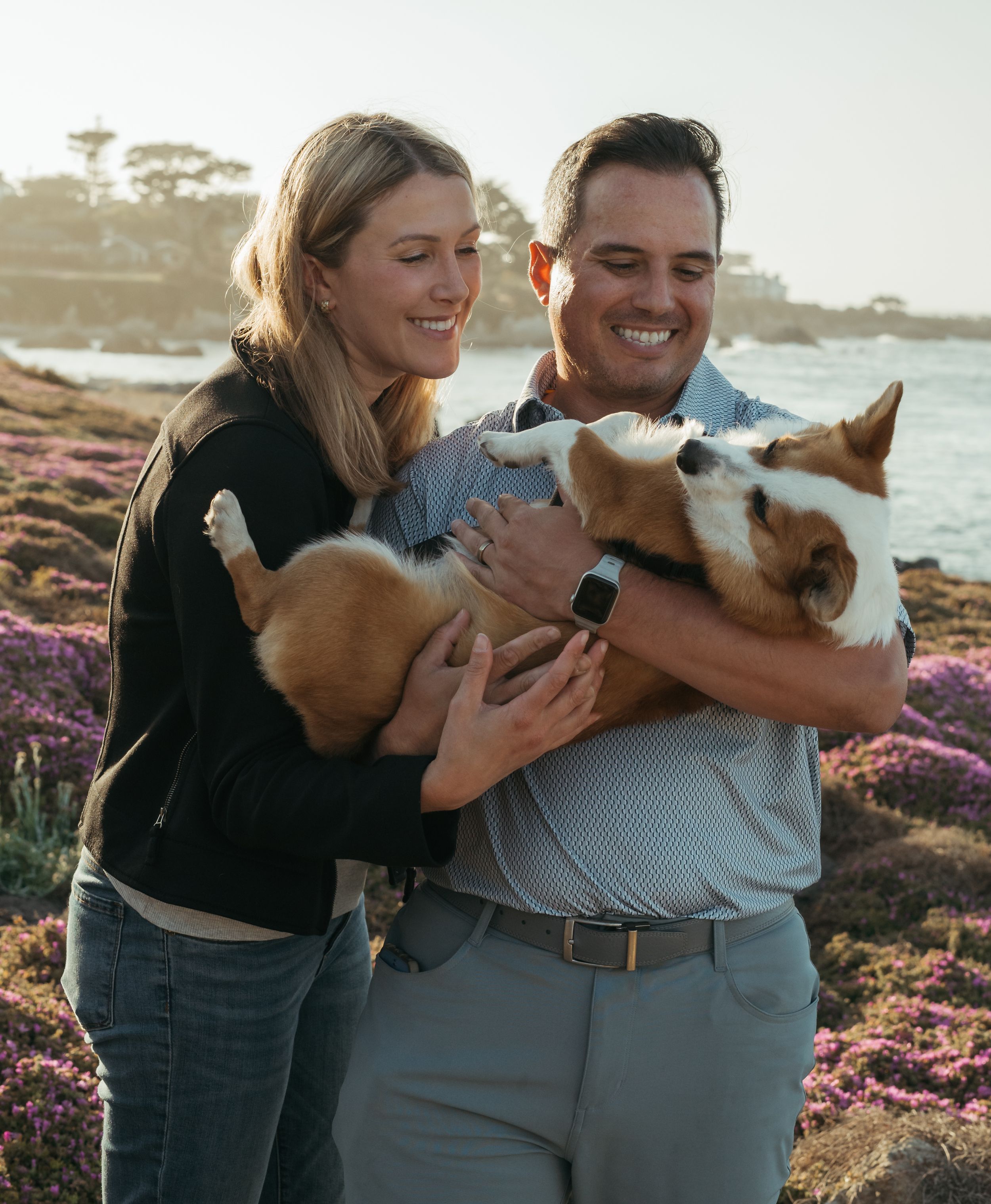 A couple shares a joyful moment at the beach while holding their playful Corgi dog among pink coastal flowers.