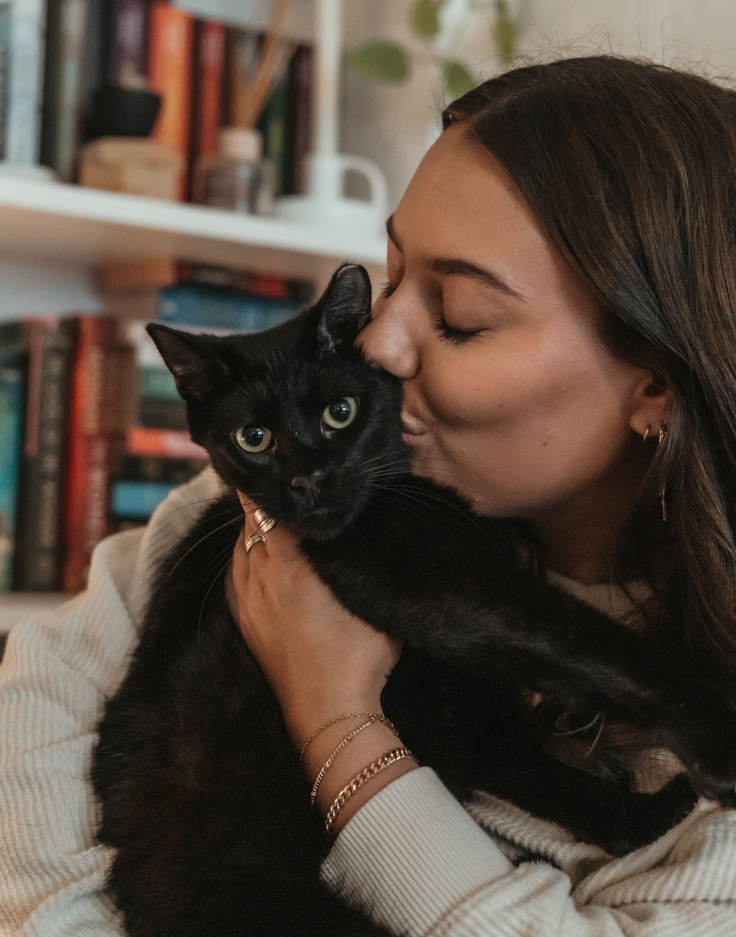 Someone in a cream sweater gives their black cat an affectionate kiss while sitting by a bookshelf.