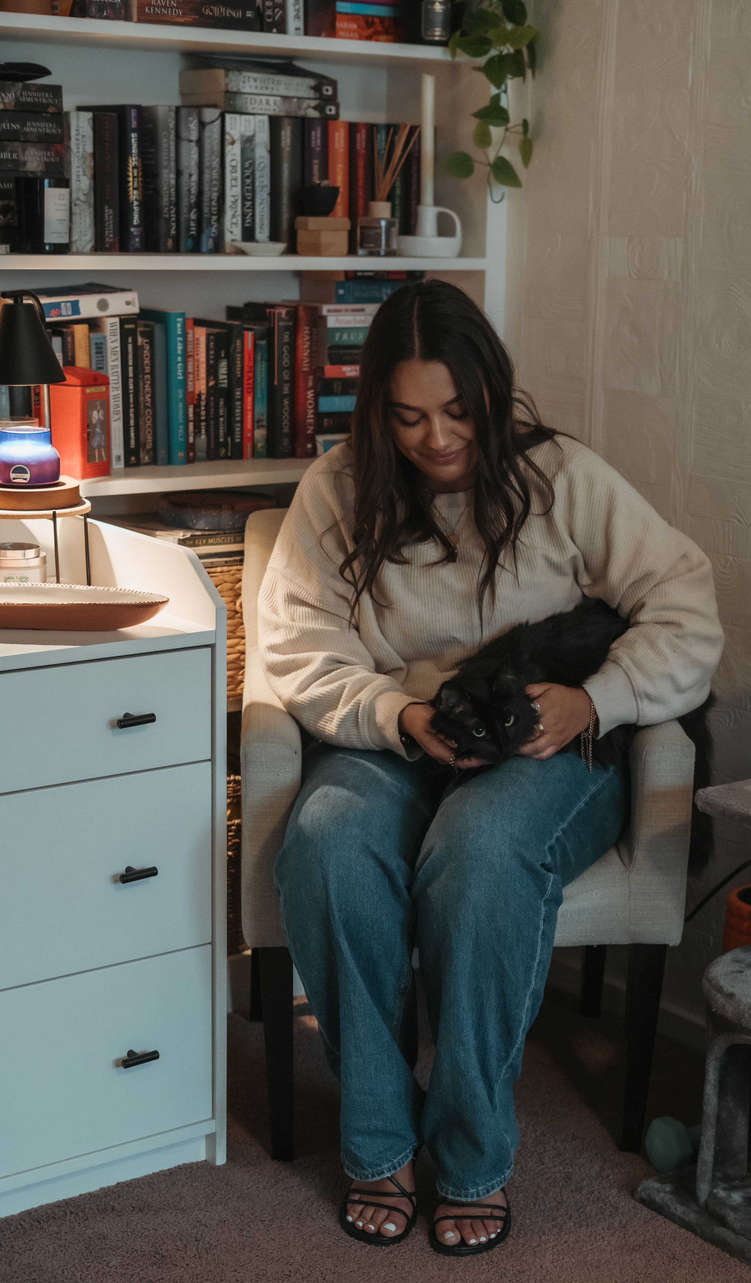 A cozy reading nook with bookshelves features someone in a cream sweater holding a black cat while sitting in a chair.