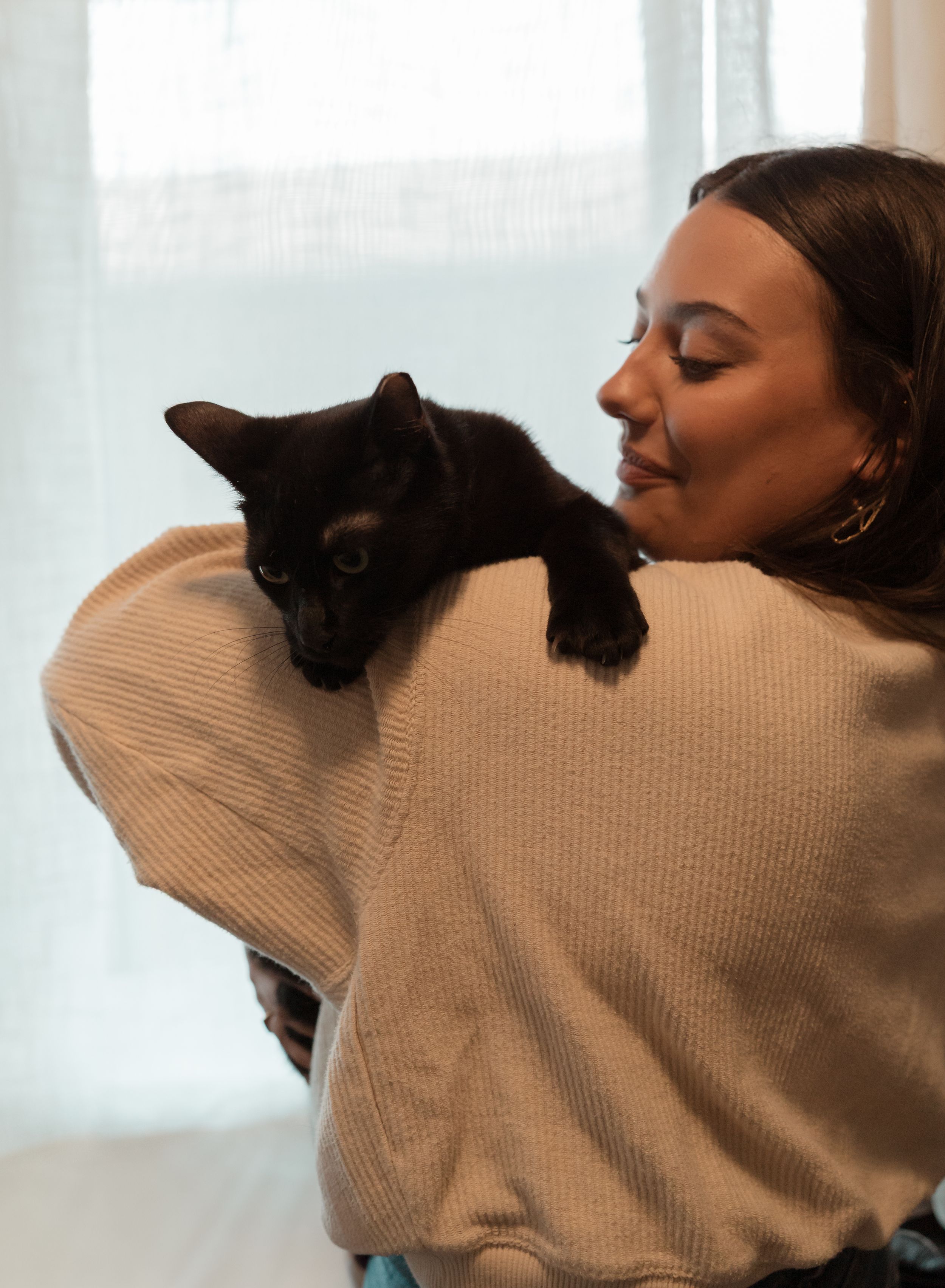 A black cat rests on someone's shoulder while they wear a beige sweater near a window.