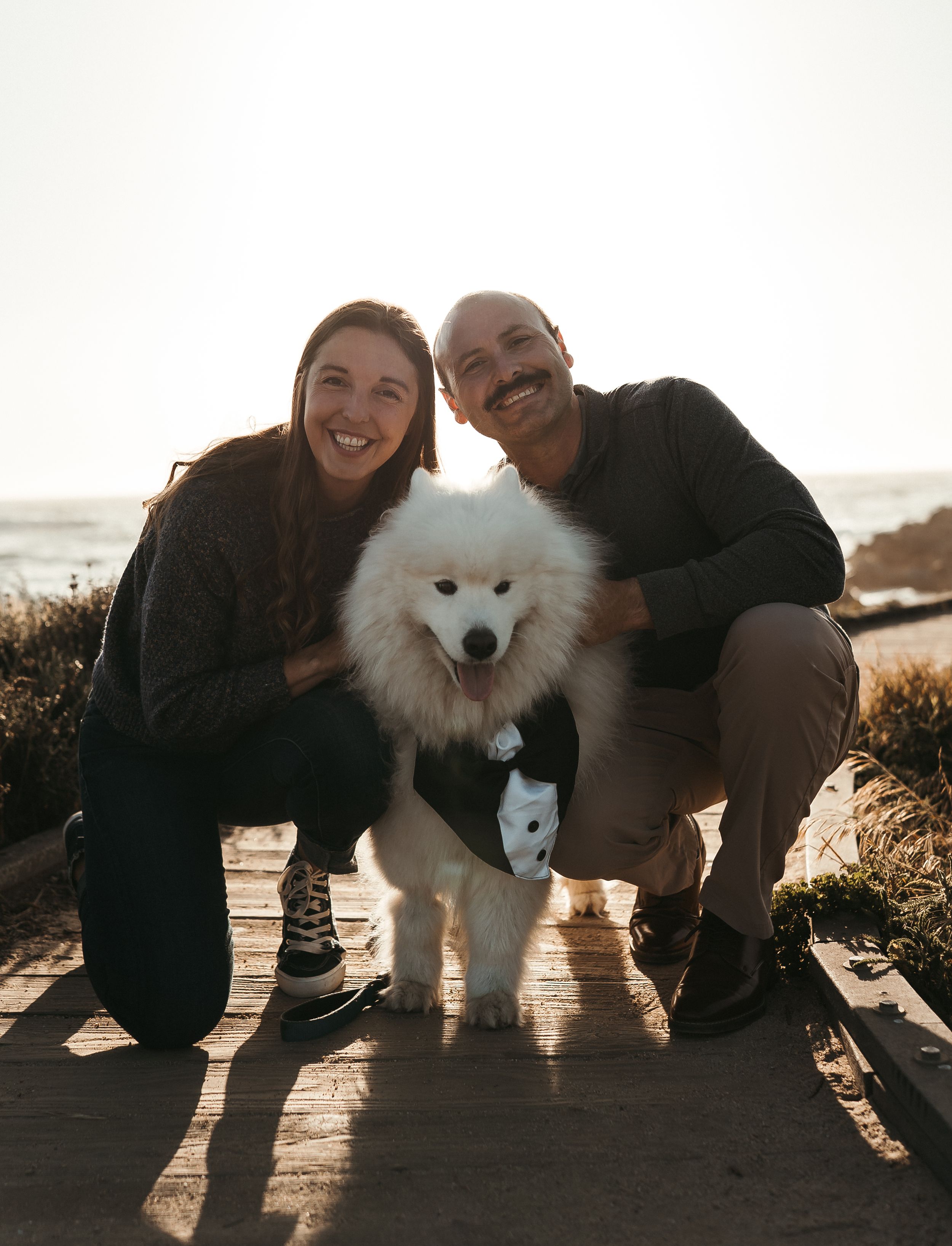 A happy couple kneeling next to their fluffy white Samoyed dog on a wooden deck at sunset.