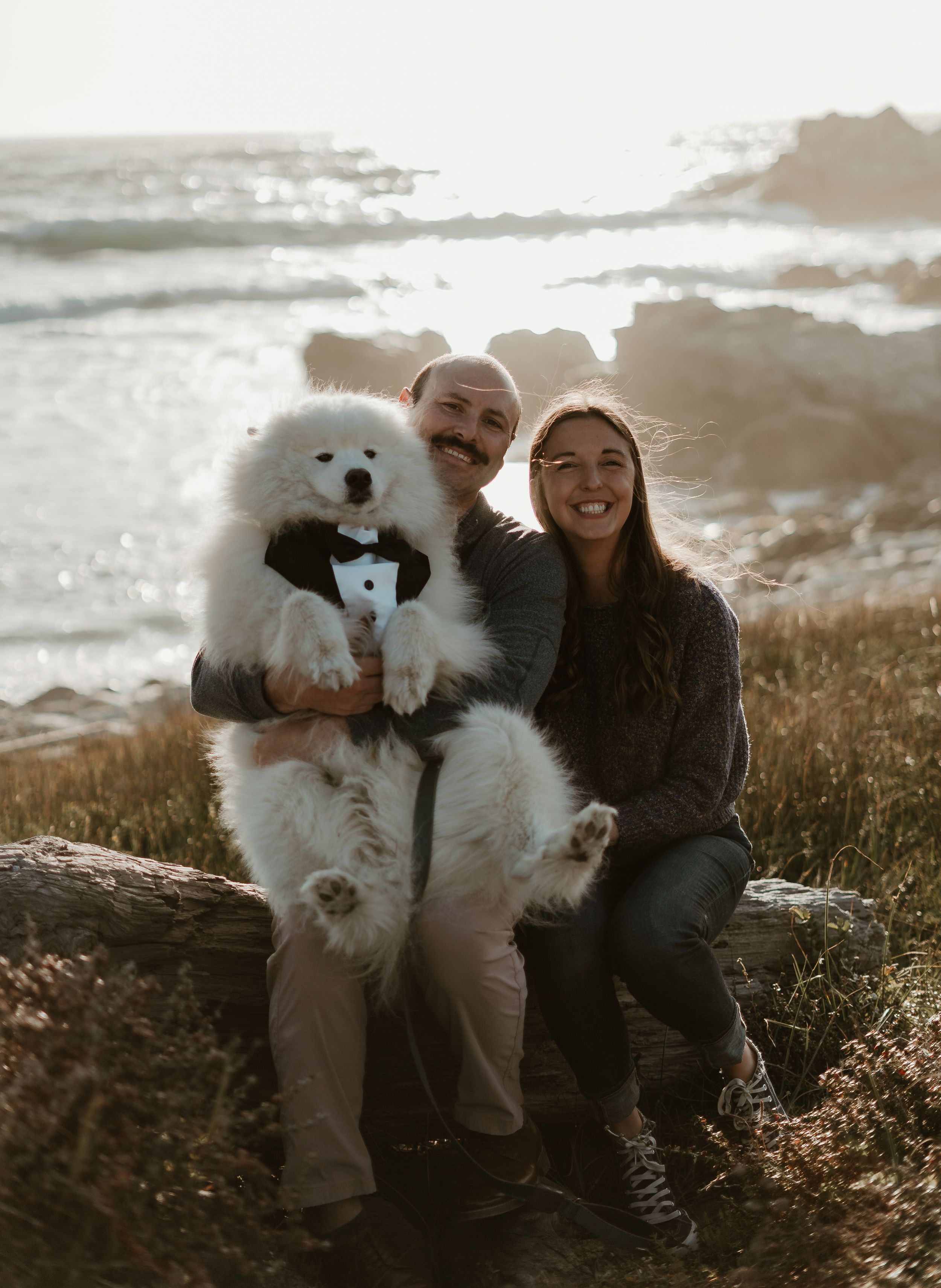A fluffy white Samoyed dog wearing a bowtie poses with two people on a scenic coastal cliff at sunset.