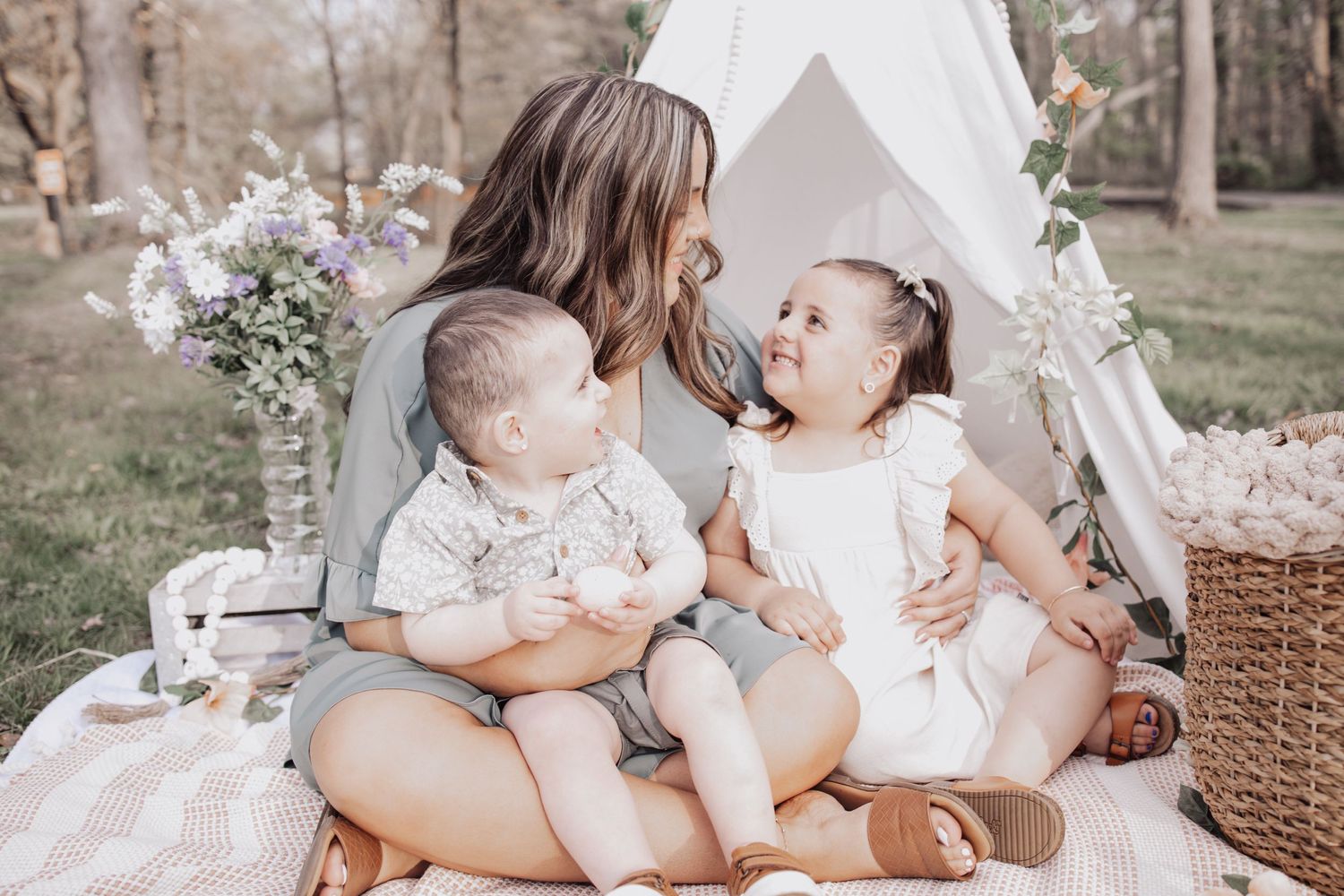 A family picnic scene with children on a blanket next to a white teepee tent with floral decorations.