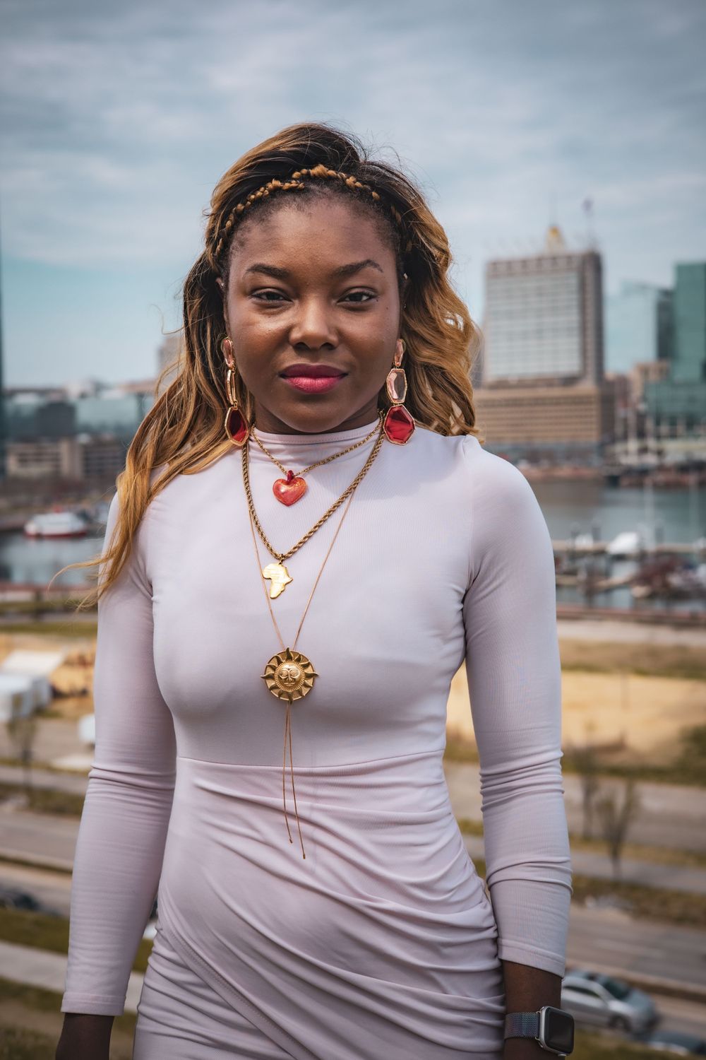 Fashionable young woman in a white long-sleeve top poses outdoors with layered necklaces against an urban backdrop.