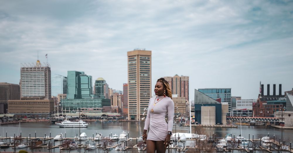 Person in winter attire stands in front of Baltimore's Inner Harbor skyline with boats and high-rise buildings in the background.