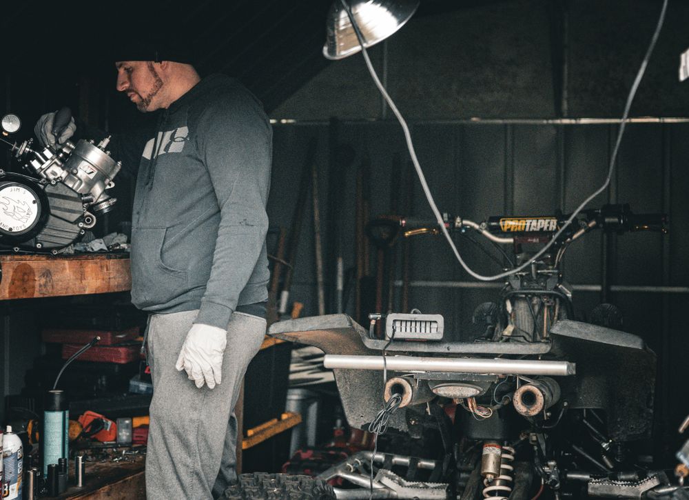 Mechanic working on motorcycle parts in dark garage with desk lamp.
