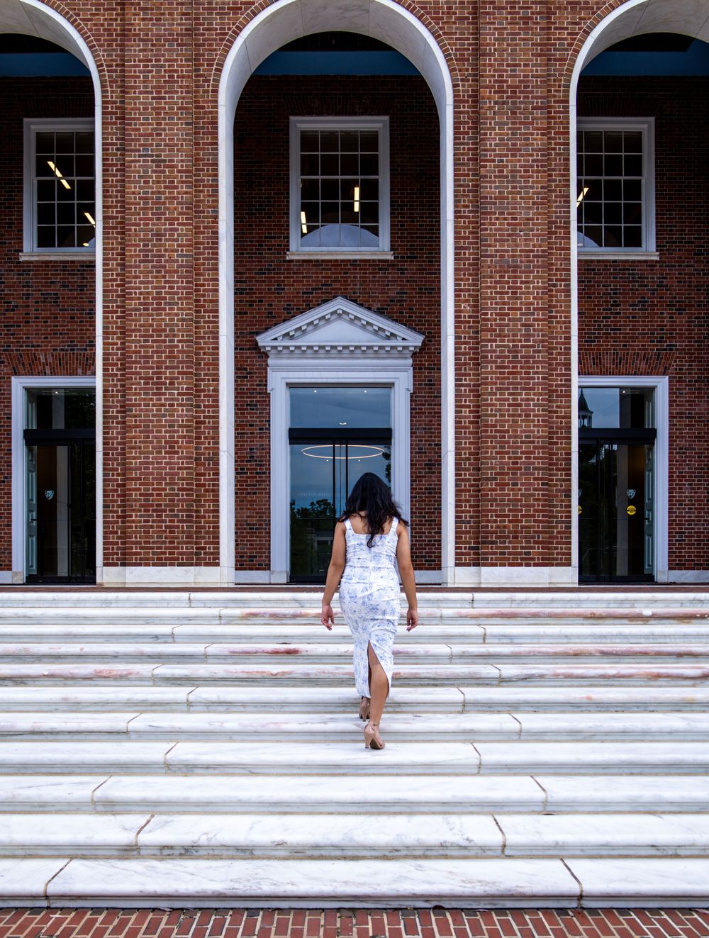 Figure in a white dress ascending grand white steps at a colonial-style brick building with arched windows and ornate doorway.