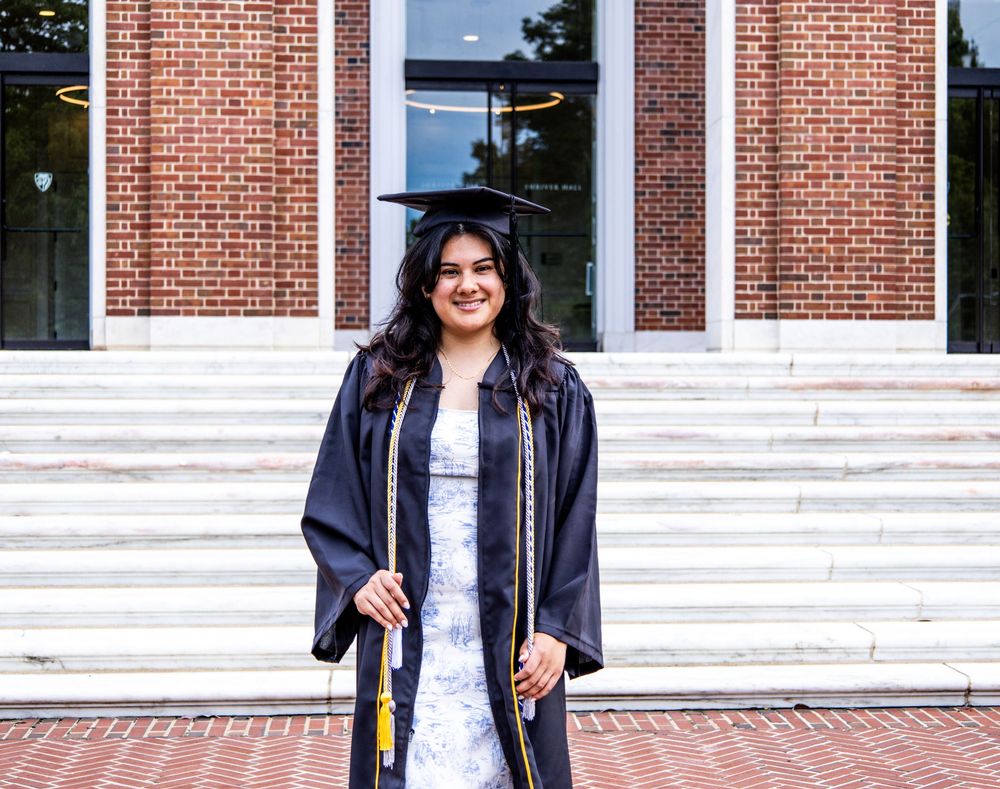 Graduate in cap and gown standing proudly on white steps in front of a brick collegiate building.