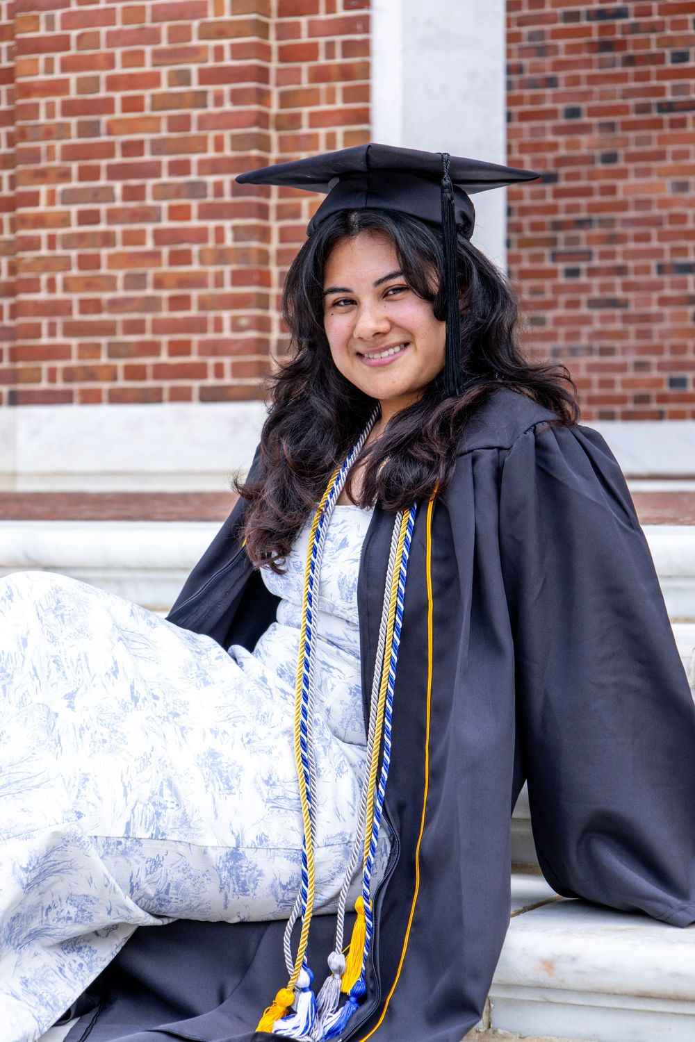 Graduate in cap and gown wearing graduation cords smiles while sitting against brick wall.