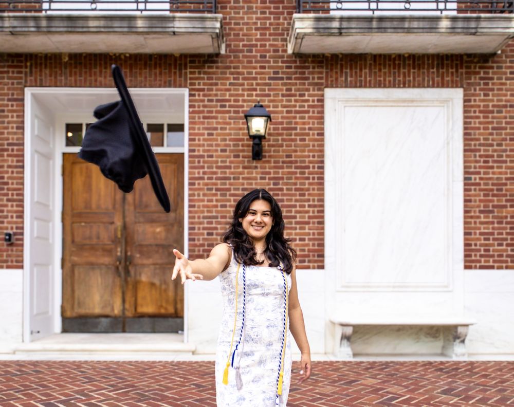 Graduate celebrates in front of a brick building while throwing their cap in the air.