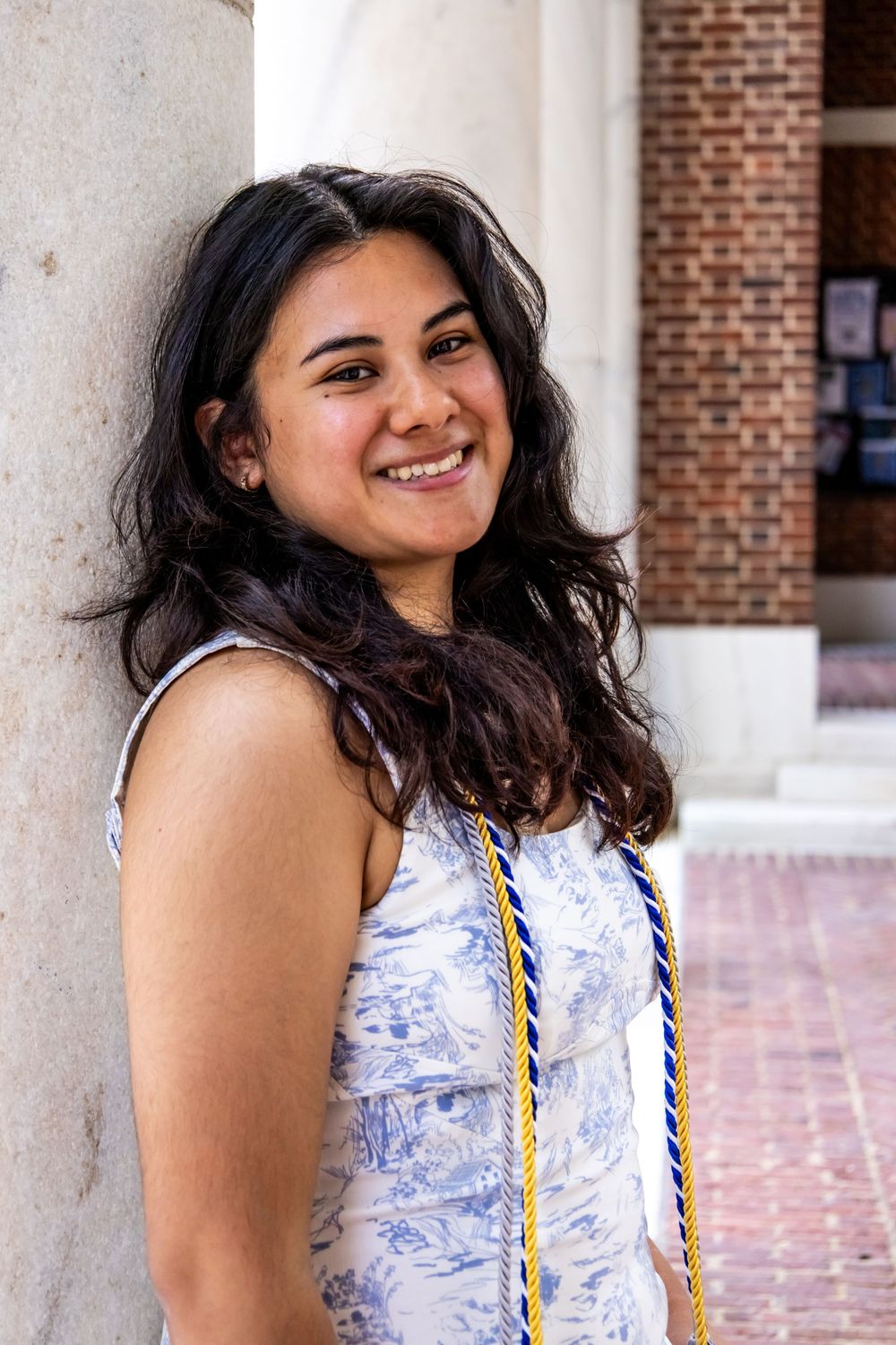 A graduate wearing a white sleeveless dress with blue details and a gold honor cord smiles outdoors by brick columns.
