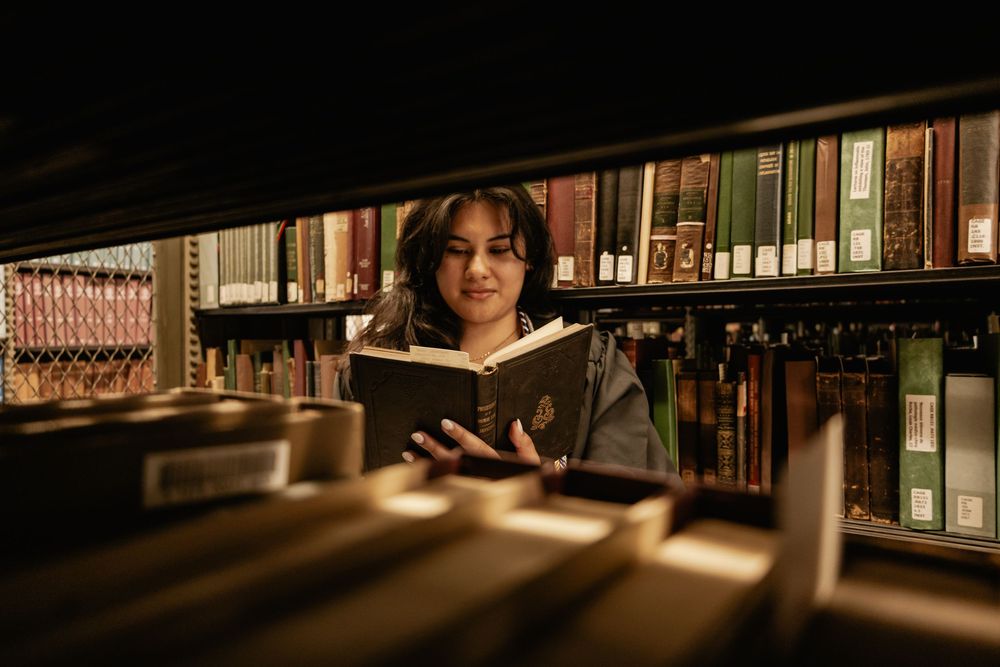 Person reading a book in a library with warm lighting and bookshelves in the background.