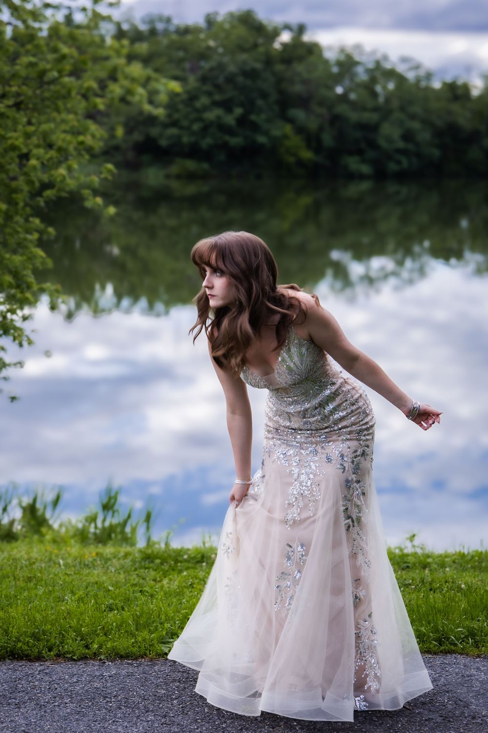 A bride in an embellished white lace gown poses gracefully by a serene lake surrounded by trees and greenery.