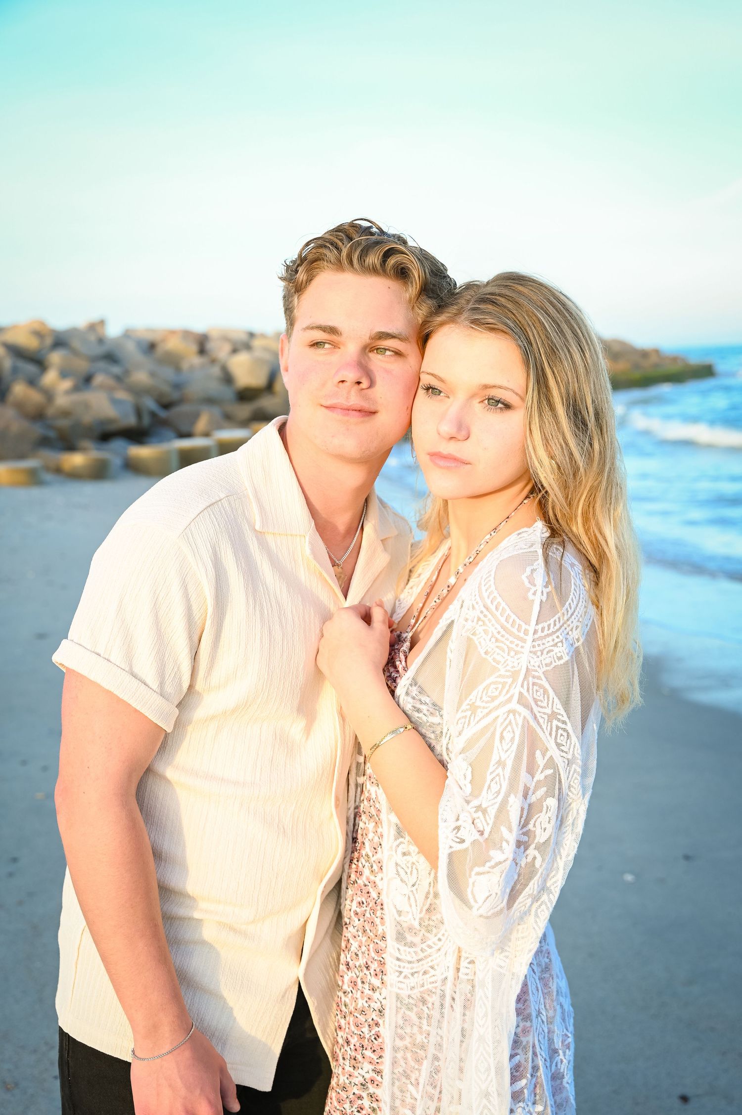 A romantic sunset beach portrait with a couple in light colored clothing against coastal rocks.