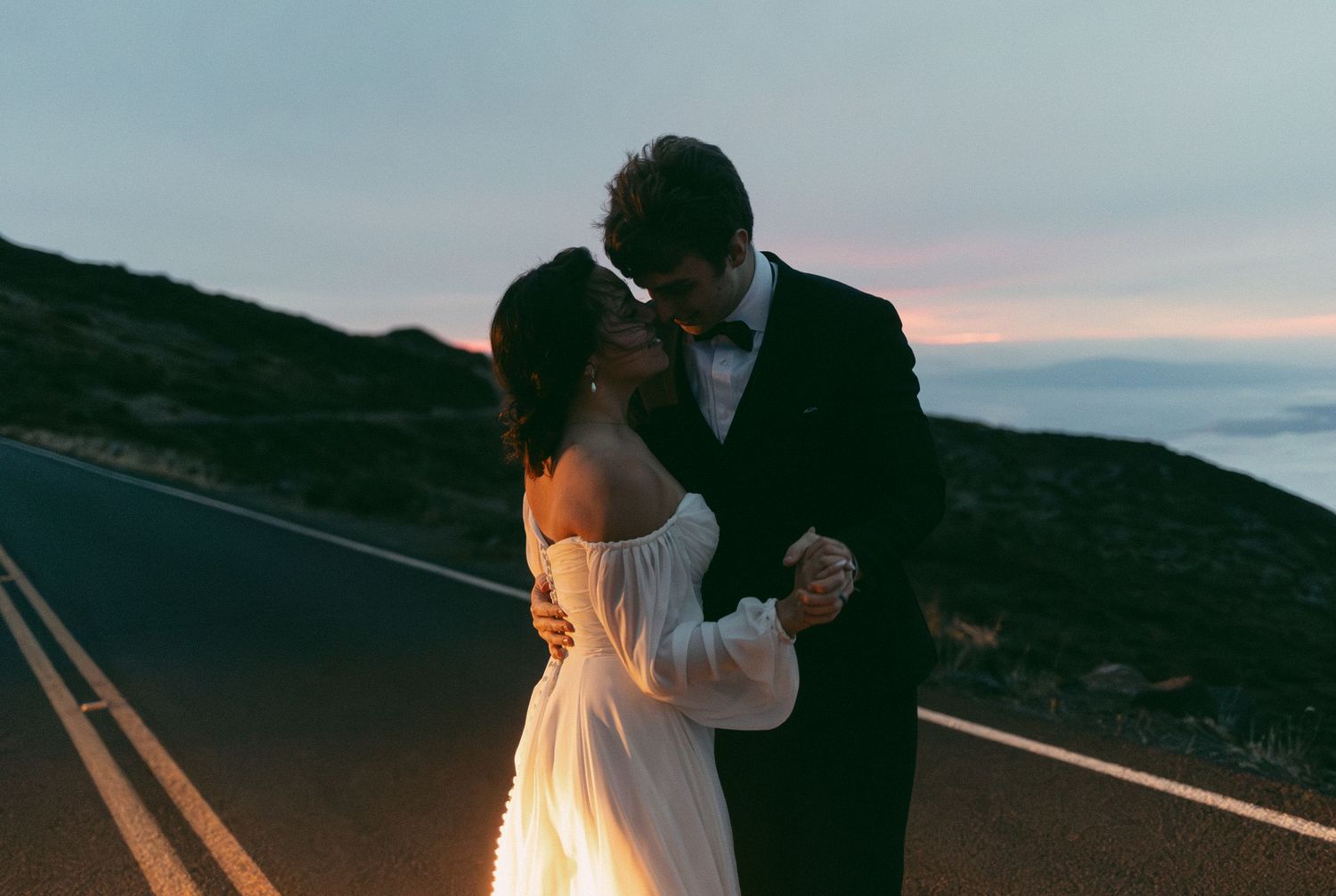 A couple in wedding attire dance together on an empty mountain road at sunset.