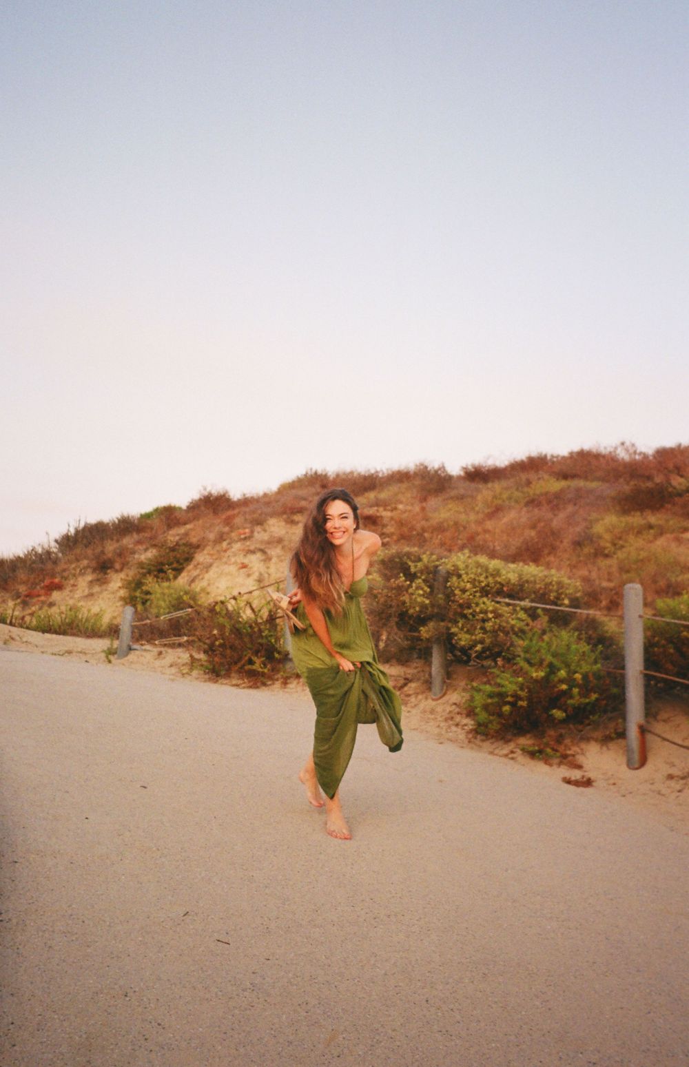 Woman in flowing green dress dances on coastal path during sunset.