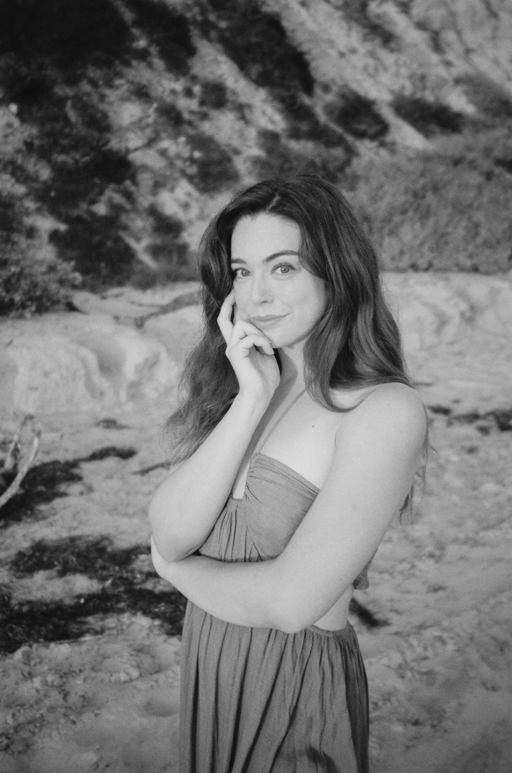 Black and white portrait of woman in dress against rocky coastal backdrop.
