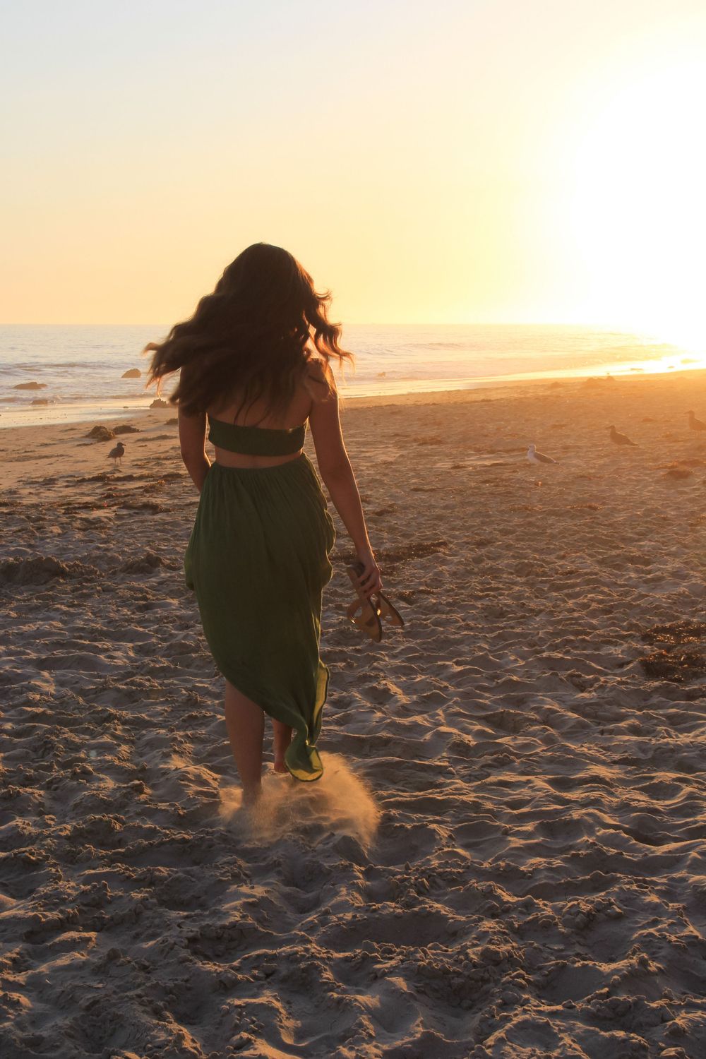 Person in green dress walks along sandy beach during golden sunset.