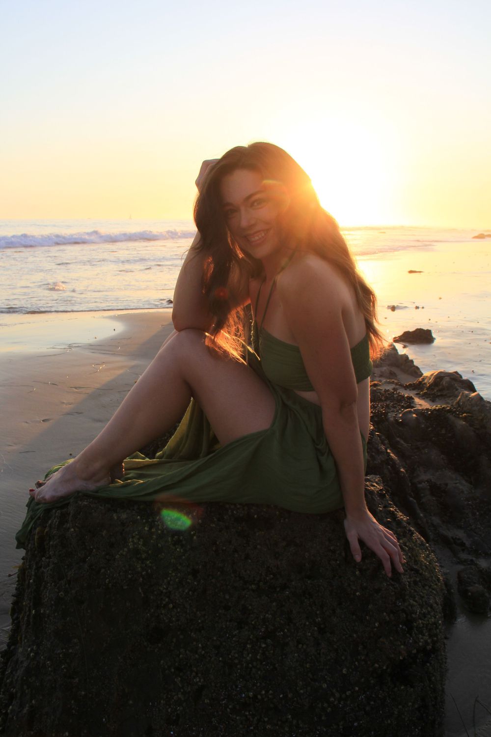Person in green dress sits on coastal rock during golden hour sunset.