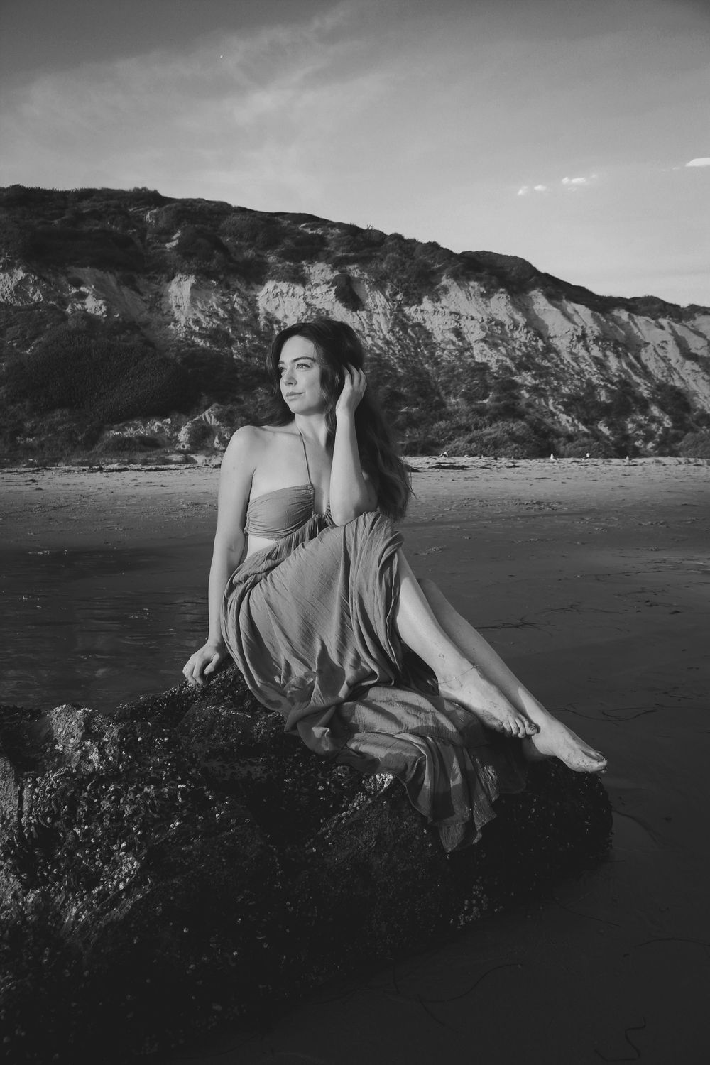 Black and white portrait of someone in flowing dress posed on beach rocks near cliffs.