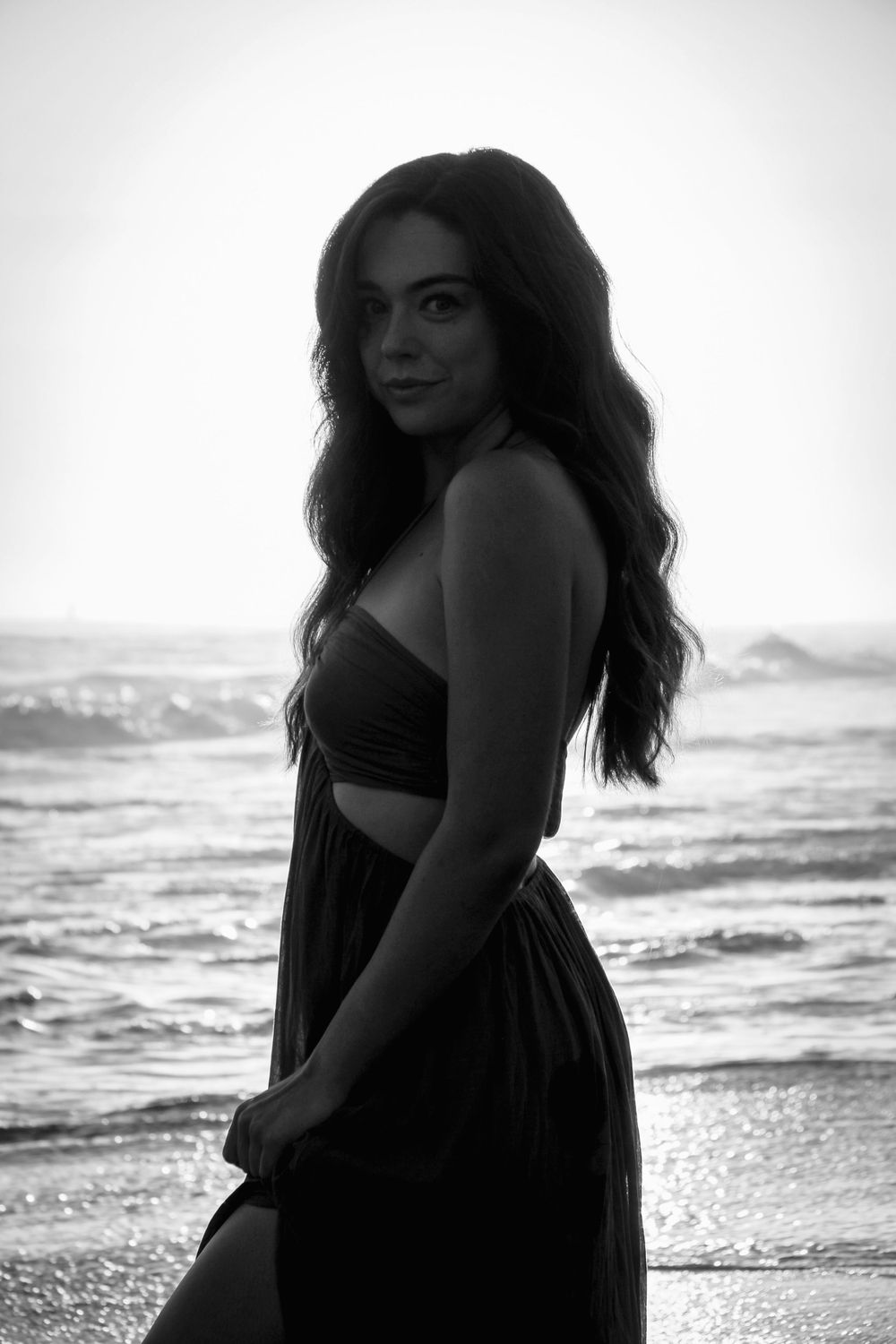 Black and white beach portrait of woman in flowing dress by ocean waves.