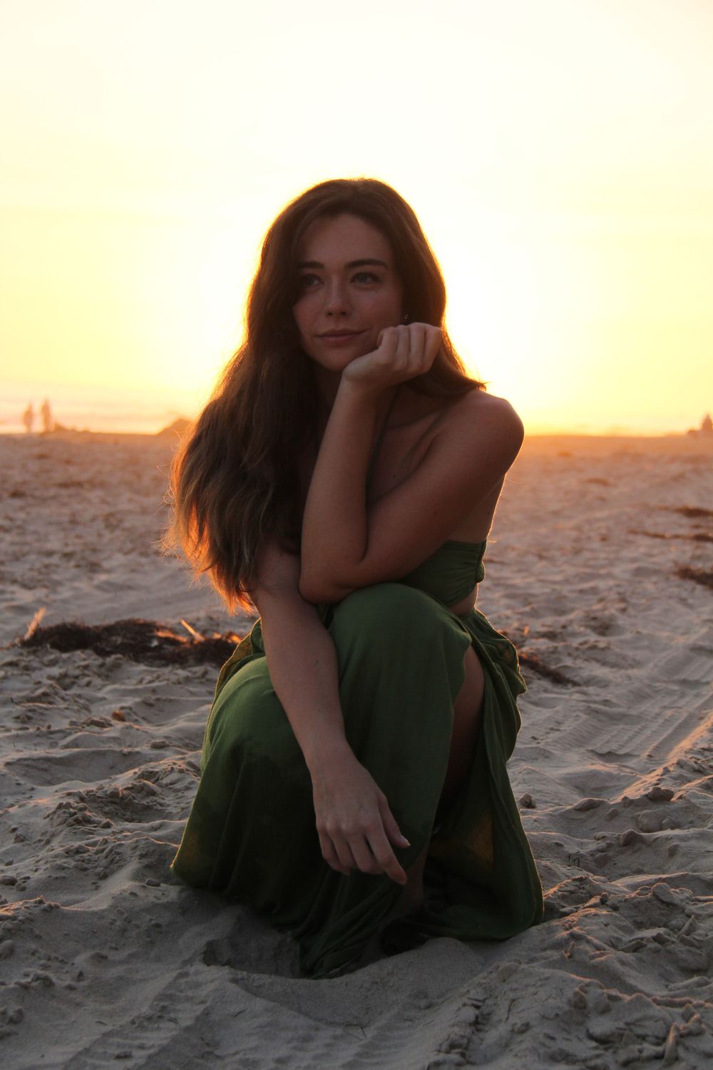 Person in green dress sits on sandy beach during golden sunset hour.